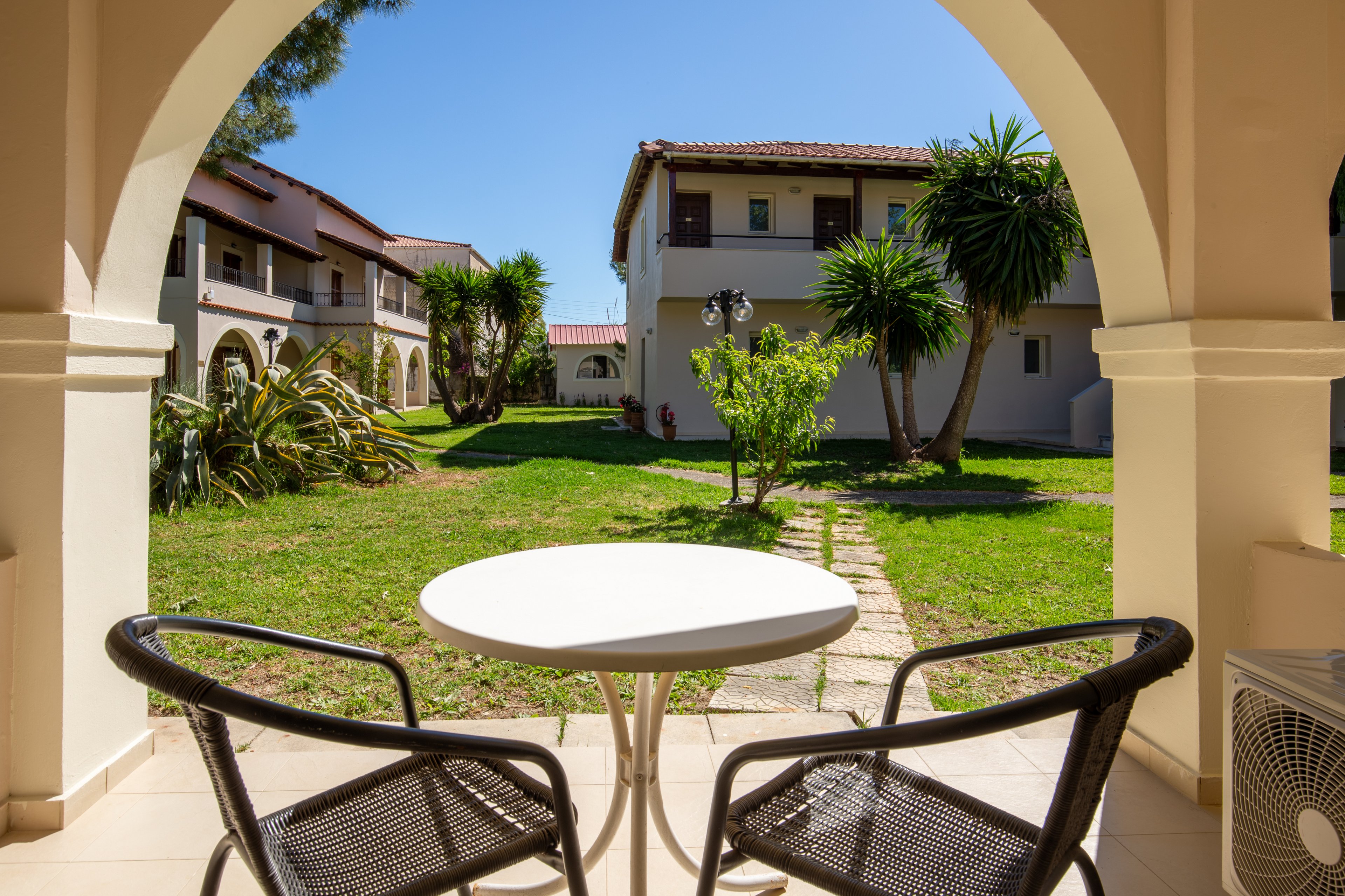 A small round table with two chairs sits on a covered terrace overlooking a sunny courtyard. The view includes well-maintained grass, tropical plants, and palm-like trees leading toward multi-story white buildings with red-tiled roofs. Arched architectural details frame the scene, creating a relaxed Mediterranean or coastal resort atmosphere. Bright daylight and clear blue sky enhance the warm, inviting vacation mood.
