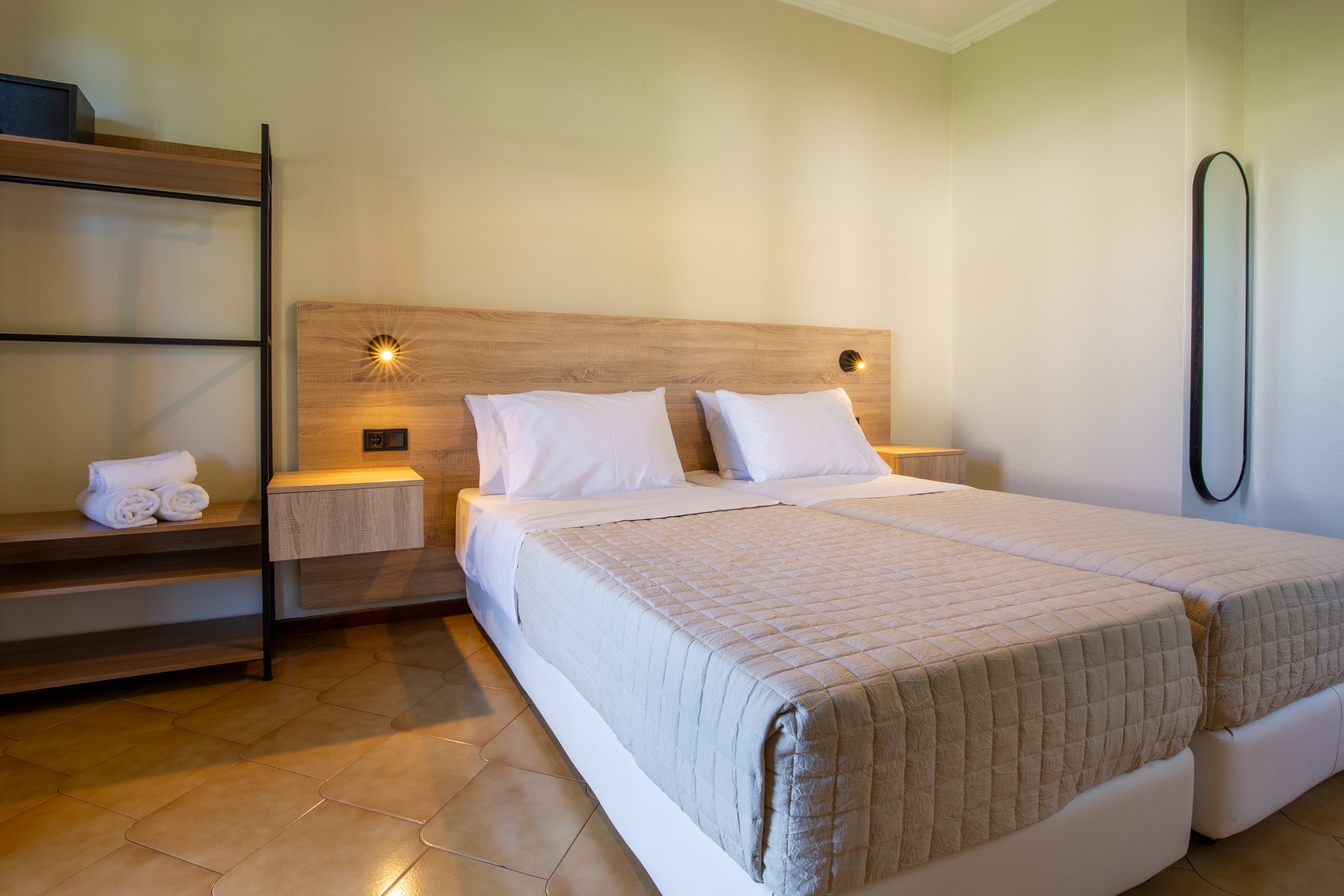 A tidy bedroom features two twin beds pushed together with beige quilted covers and white pillows. Wall-mounted bedside tables and small round lights create a simple, contemporary look. An open shelving unit with neatly folded white towels stands to the left, while a tall oval mirror is mounted on the right wall. The room is warmly lit, with neutral tones and clean lines emphasizing a minimalist, comfortable atmosphere.