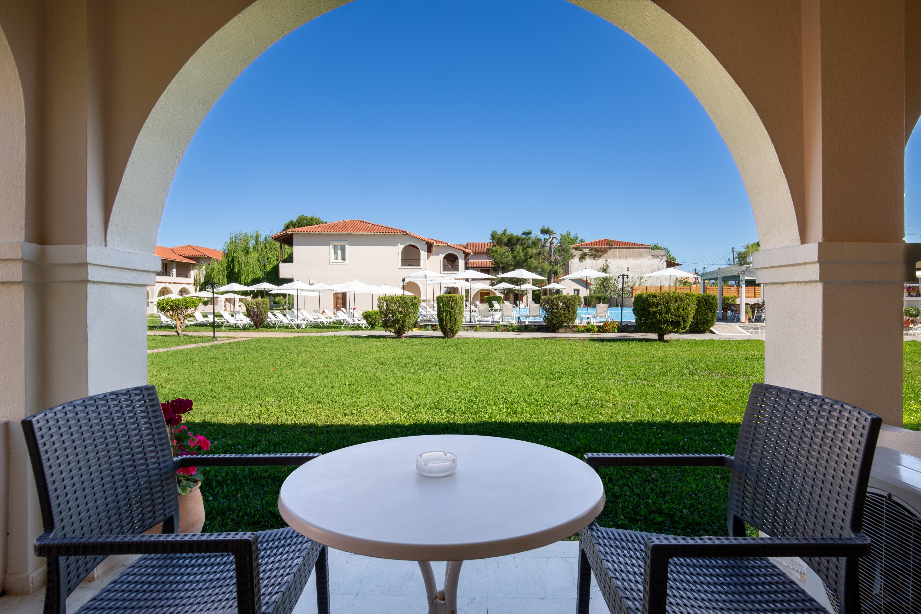 A small round table with two chairs sits on a covered terrace framed by an archway. Beyond the terrace, a manicured green lawn leads to a resort complex with white buildings and red-tiled roofs. Sun loungers and umbrellas surround a bright blue swimming pool in the center of the scene. The sky is clear and vivid blue, creating a calm and inviting vacation atmosphere.
