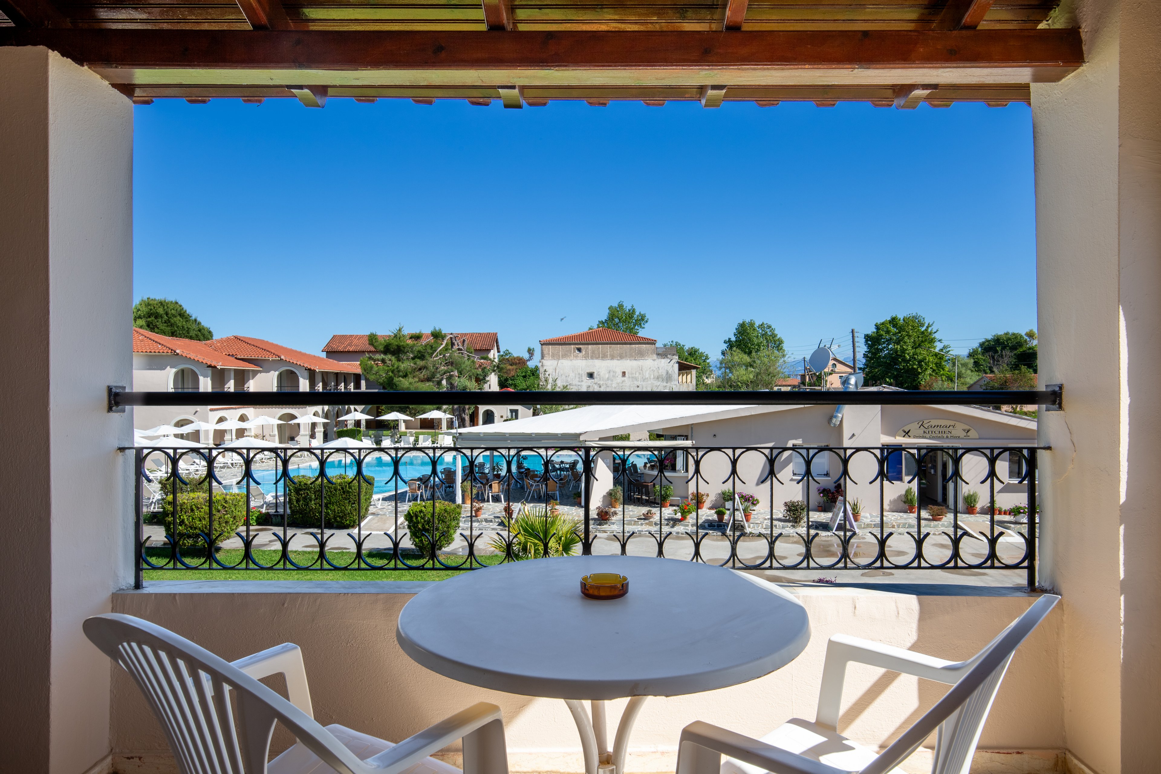 A small balcony with a round table and two white chairs overlooks a bright resort courtyard. Beyond the decorative railing, a large outdoor swimming pool is surrounded by sunlit buildings and greenery. The sky is clear and blue, creating a relaxed vacation atmosphere. The scene suggests a peaceful place to sit and enjoy the view.