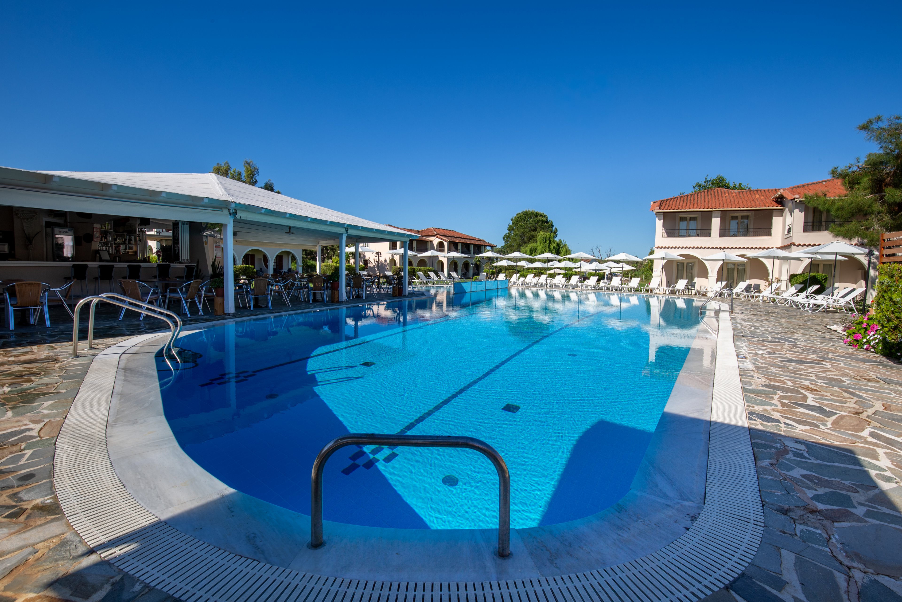 An outdoor swimming pool is featured at a sunny resort with clear blue water and metal handrails in the foreground. White sun loungers line the poolside terrace, facing the water under a bright blue sky. A covered patio area with tables and chairs sits to the left, while low-rise buildings with red-tiled roofs frame the background. The scene suggests a relaxing vacation atmosphere in a warm, coastal destination.