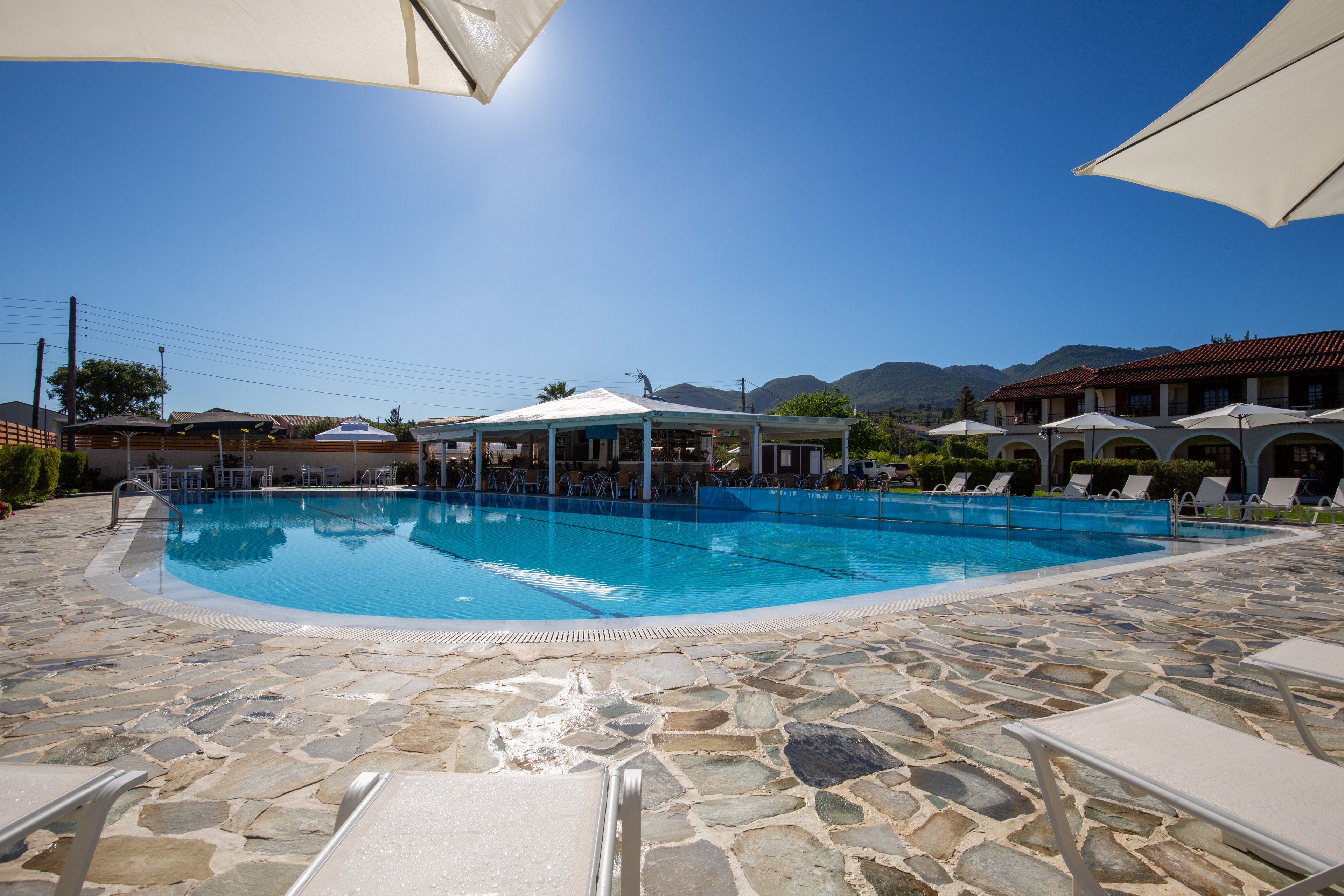 A clear blue swimming pool is surrounded by stone paving and white sun loungers at a resort. White umbrellas provide shade around the pool area under a bright, cloudless sky. In the background, low-rise buildings with tiled roofs and arched terraces frame the scene. Distant mountains and greenery add a scenic backdrop, creating a relaxed holiday atmosphere.