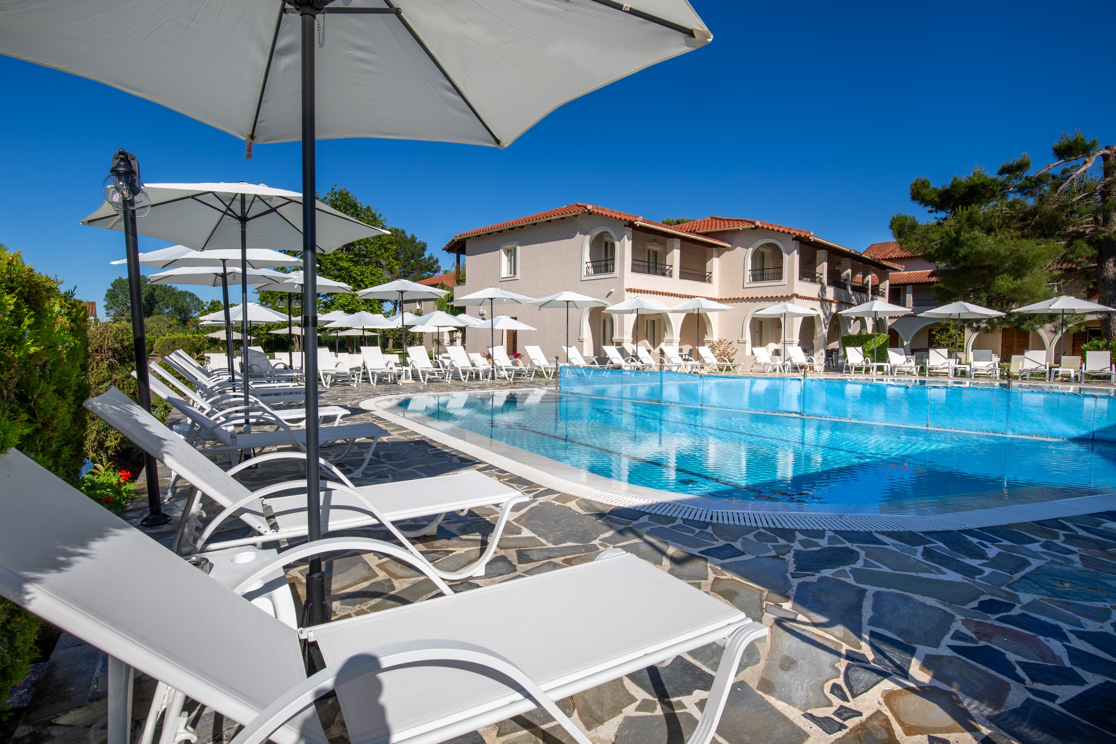 A bright outdoor swimming pool is surrounded by white lounge chairs and umbrellas at a resort. The pool area is paved with patterned stone tiles and bordered by green shrubs. A Mediterranean-style building with balconies and red-tiled roofs stands in the background under a clear blue sky. The scene suggests a relaxing vacation atmosphere in warm weather.