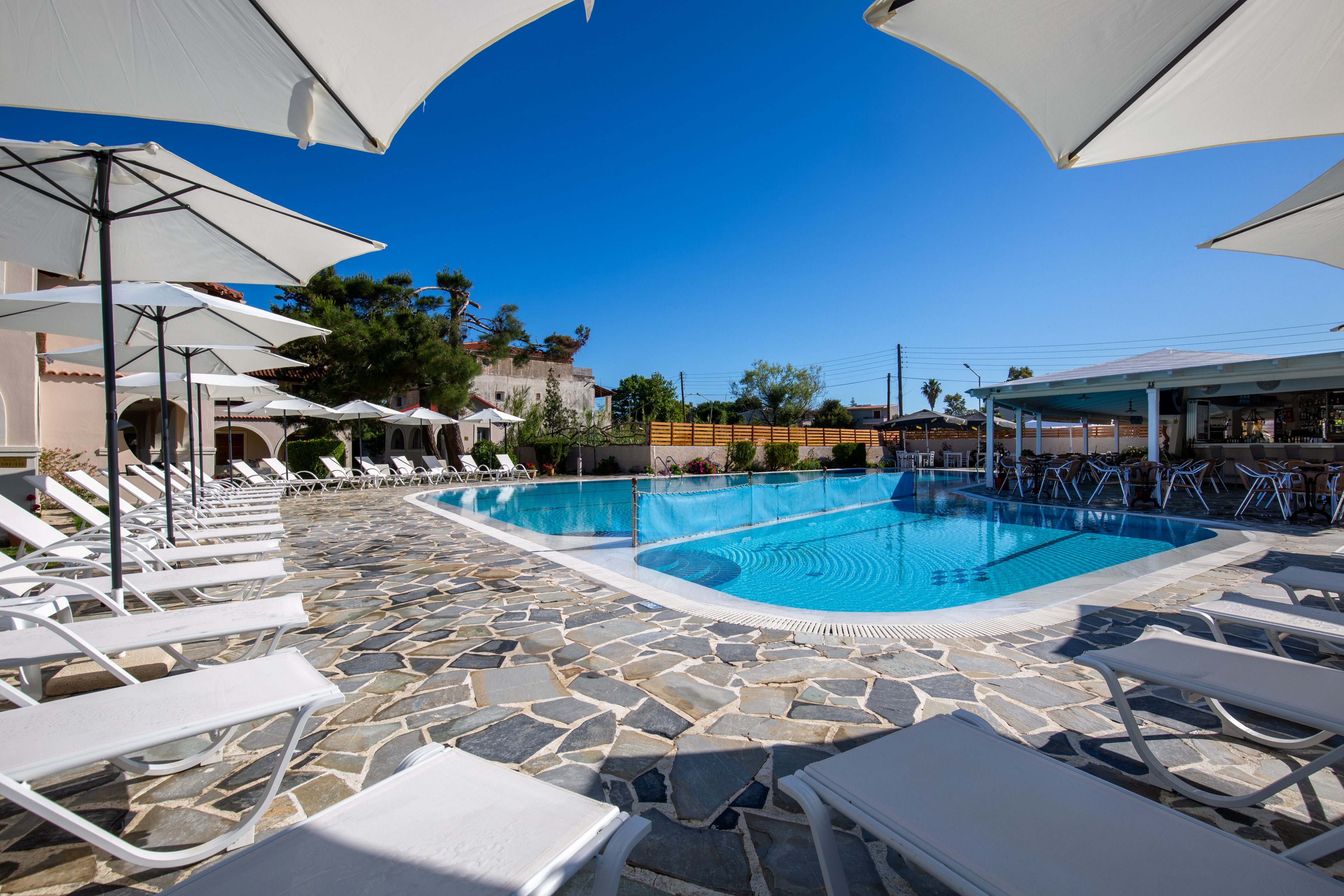 An outdoor resort swimming pool is surrounded by white sun loungers and large umbrellas on a stone-tiled deck. The clear blue water reflects the bright sky, creating a relaxing vacation atmosphere. Buildings and a poolside bar or café are visible in the background, suggesting guest amenities nearby. The scene is empty of people, emphasizing tranquility and leisure under strong daylight.