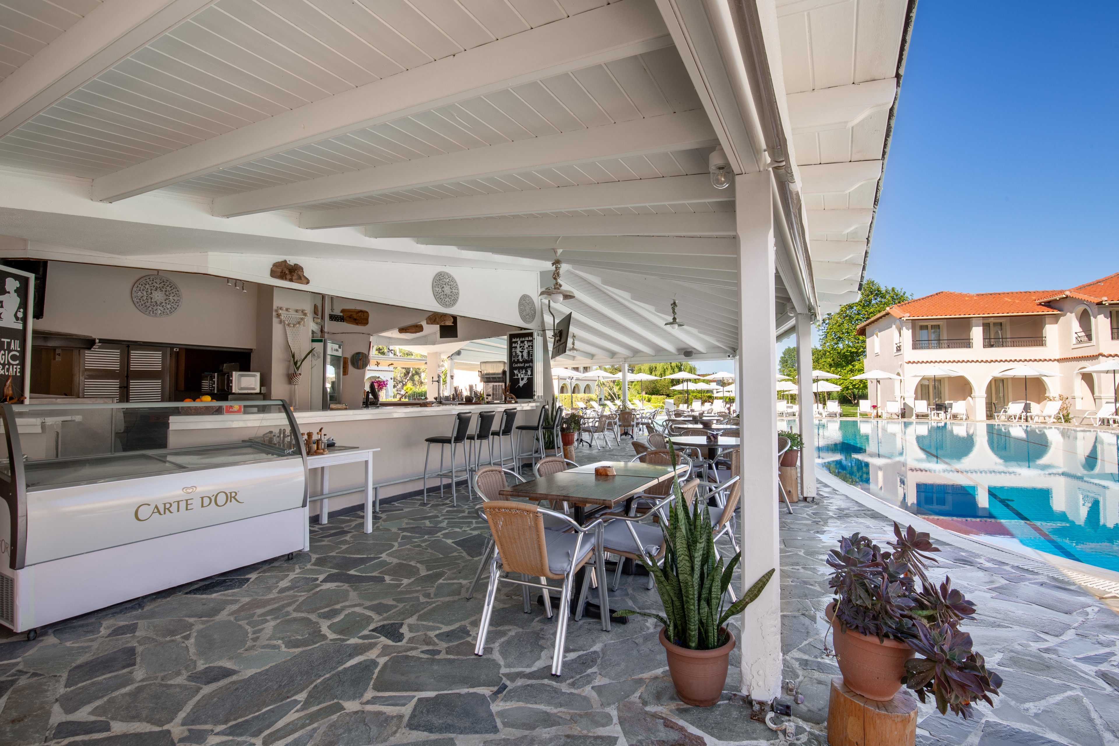 An open-air café sits beside a clear blue swimming pool at a sunny resort. Metal and wicker chairs surround tables on a stone-tiled floor under a white wooden roof. A refrigerated display counter with the text "Carte D'Or" is visible near the bar area. In the background, sun loungers, umbrellas, and a multi-story building with red roof tiles complete the relaxed vacation setting.
