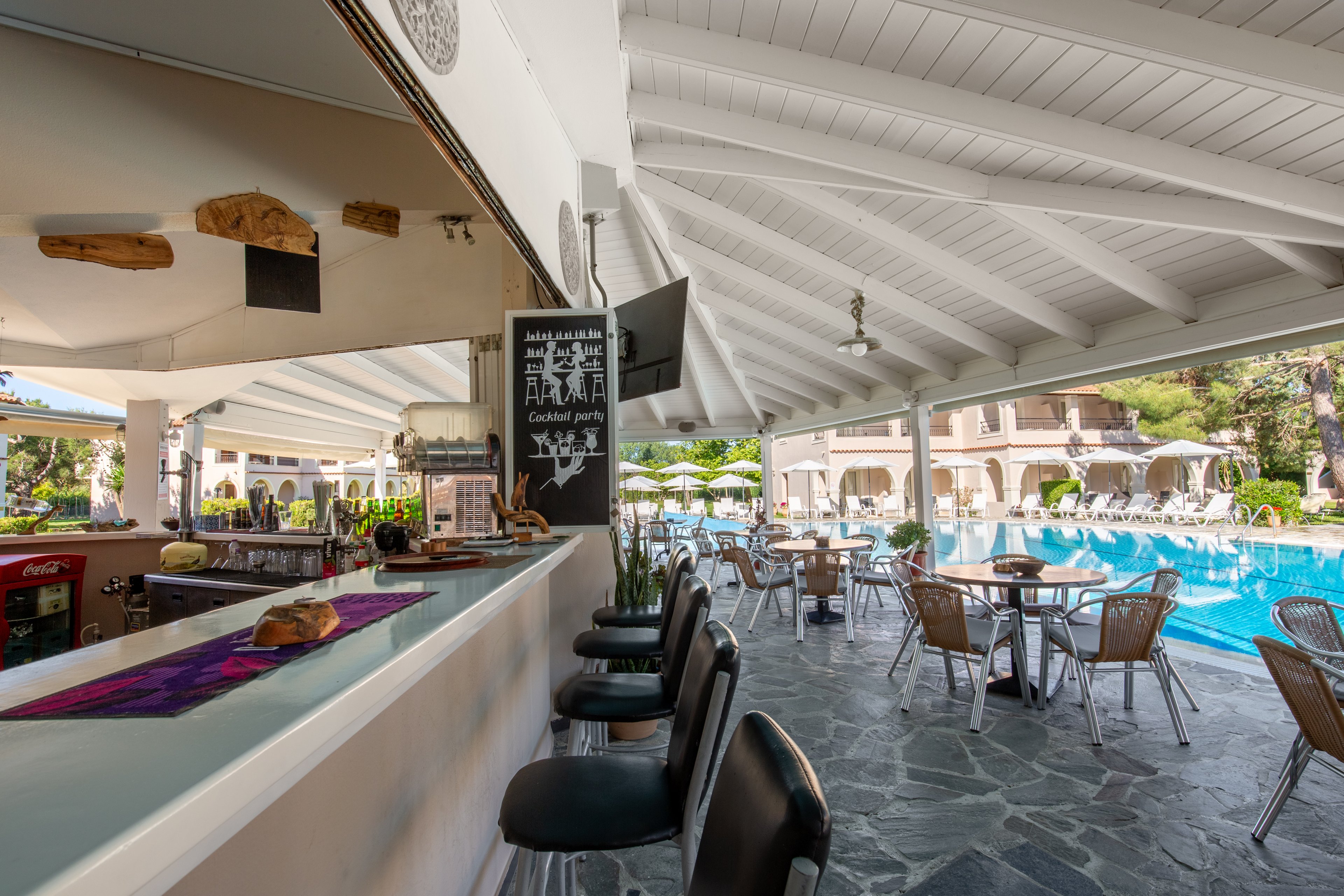 An open-air bar overlooks a resort swimming pool with tables and chairs arranged for outdoor dining. The covered terrace features a white wooden ceiling, stone flooring, and bar stools along the counter. A chalkboard menu and various food items are displayed on the bar, while sun loungers and umbrellas line the poolside in the background. Lush greenery and multi-story resort buildings frame the relaxing holiday setting.