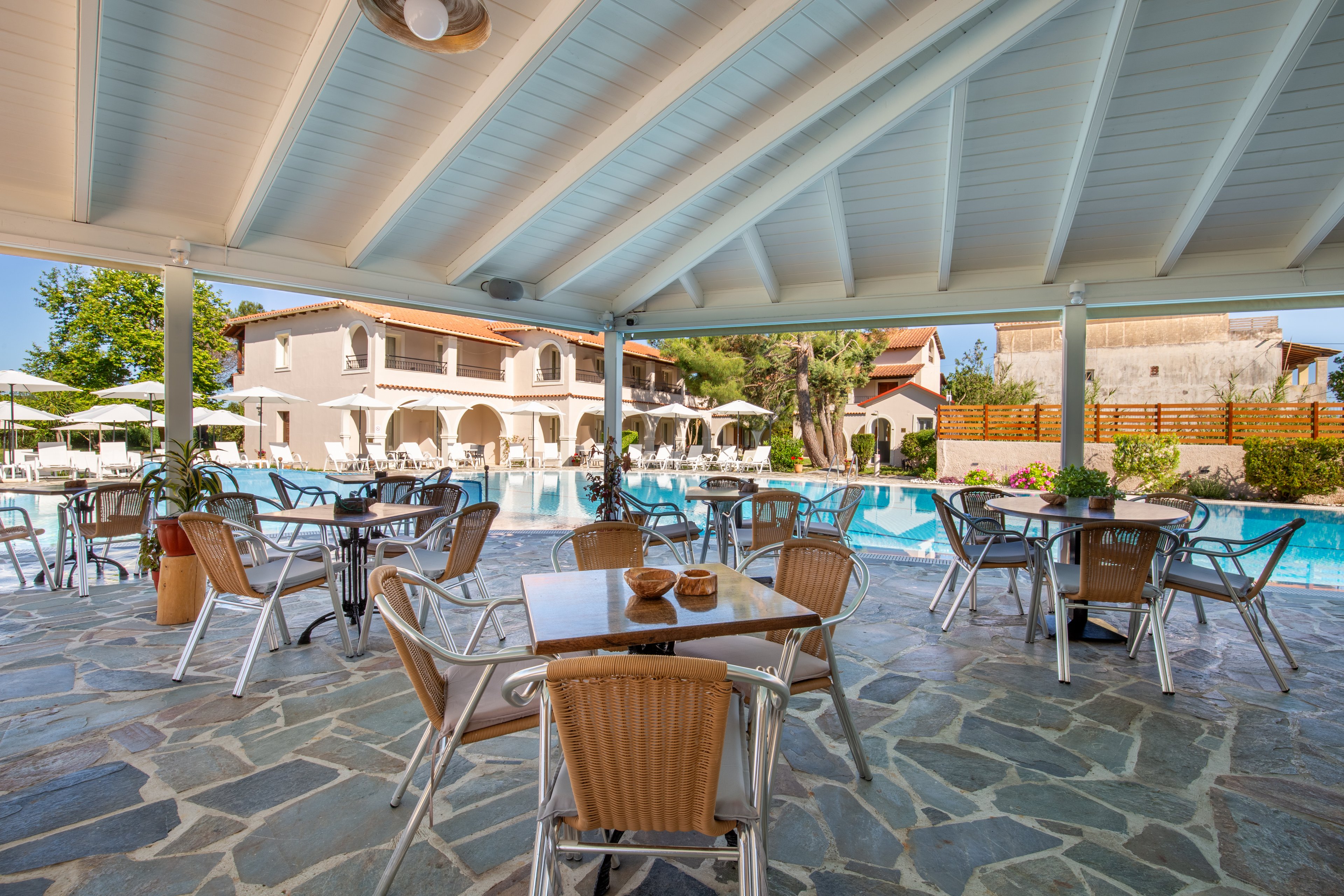 Open-air seating is arranged under a covered terrace beside a clear blue swimming pool. Metal and wicker chairs surround small tables on a stone-paved floor, creating a relaxed dining area. In the background, a Mediterranean-style building with arches and balconies faces the pool, along with sun loungers and umbrellas. Bright daylight and lush greenery contribute to a calm, vacation atmosphere.