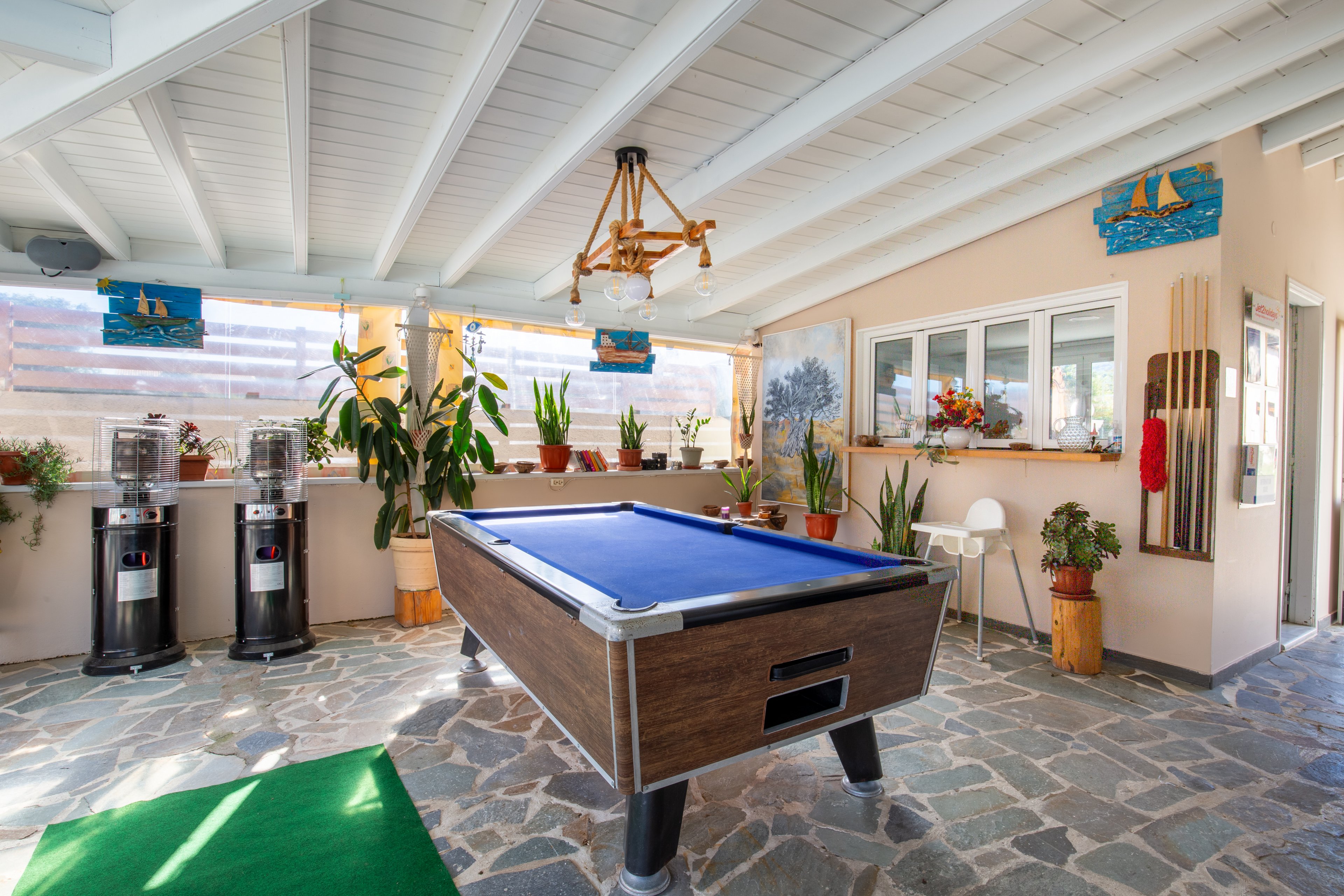 A blue felt pool table stands in the center of a bright indoor recreation room. The space features stone flooring, white wooden ceiling beams, and large windows letting in natural light. Potted plants, wall art, and decorative items line the walls and window ledge, creating a cozy atmosphere. Two water dispensers and a small seating area with a white chair are visible in the background.
