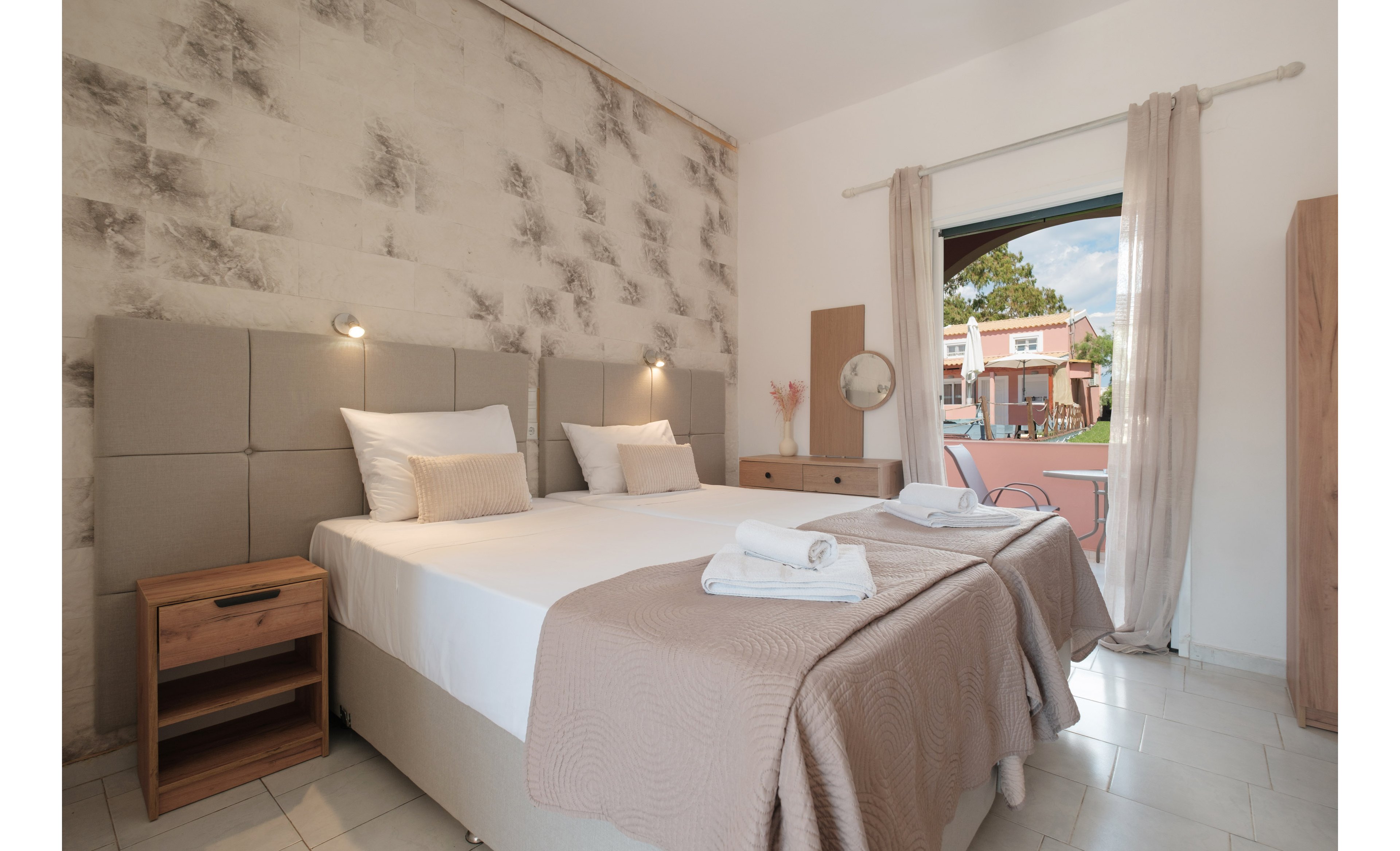 A neatly arranged hotel bedroom features two twin beds pushed together with beige headboards and matching bedspreads. Soft lighting and neutral tones create a calm, inviting atmosphere. A wooden nightstand and dresser add warmth, while sheer curtains frame a large open doorway. Outside, a patio area and neighboring buildings are visible under a partly cloudy sky.