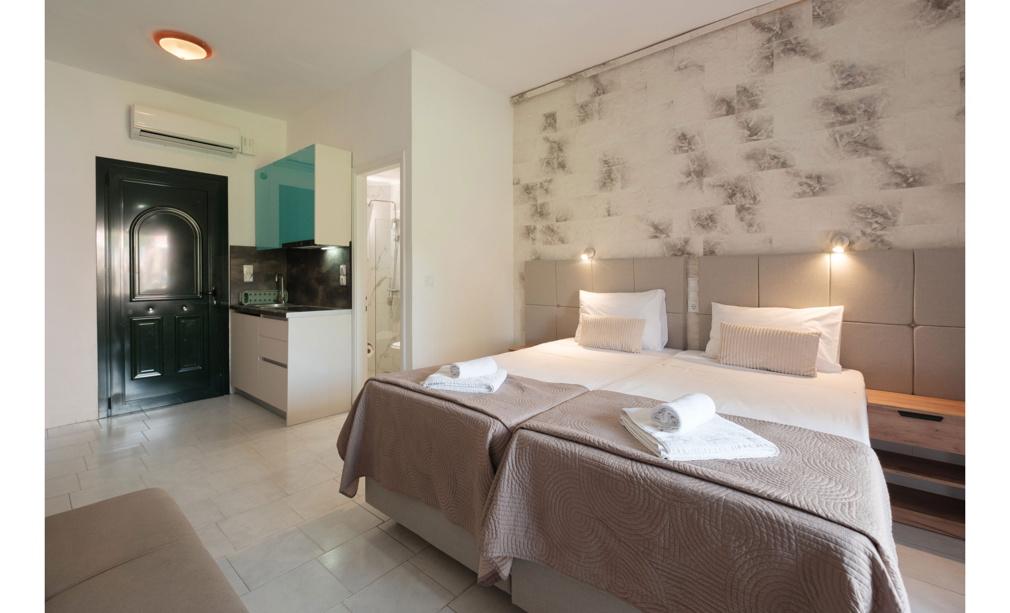 A contemporary hotel room features two neatly made twin beds with folded towels placed on top. The space includes a small kitchenette area and a partially open bathroom door in the background. Soft wall-mounted lights and a textured accent wall create a warm, inviting atmosphere. Neutral tones and minimal decor give the room a clean, modern look suitable for travelers.