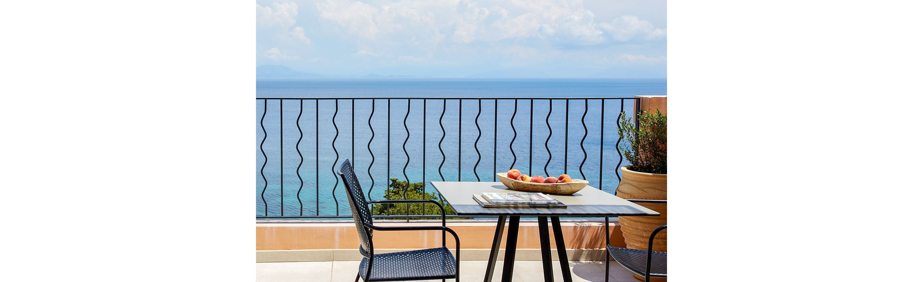 A modern outdoor terrace overlooks a calm blue sea beyond a decorative metal railing. In the foreground, a square table holds a shallow bowl filled with apples and a stack of magazines. A black mesh chair sits beside the table, suggesting a relaxed seating area. Large planters with greenery frame the scene, adding warmth and a Mediterranean feel.