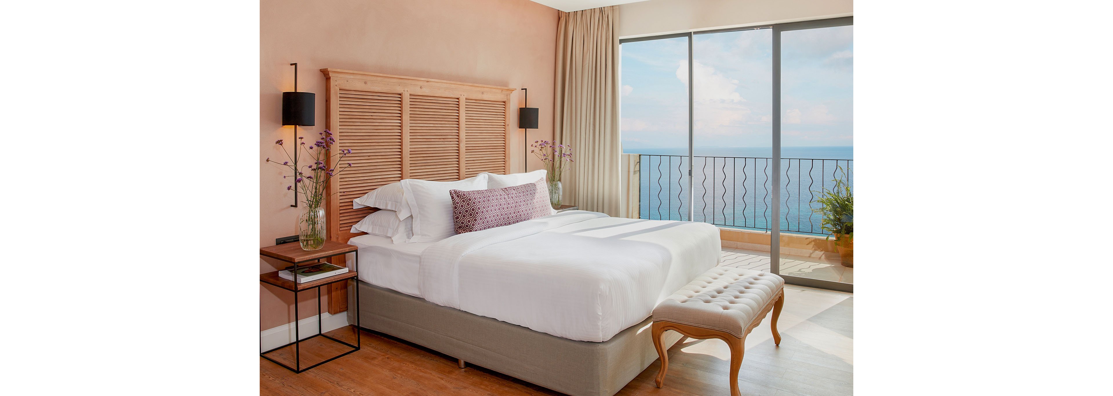 A neatly made double bed with white linens sits in a stylish hotel bedroom. The room features a wooden headboard, bedside tables with flowers, and wall-mounted lamps against soft peach walls. Large floor-to-ceiling windows open to a balcony with a wrought-iron railing overlooking the sea. A tufted bench at the foot of the bed and light curtains add to the airy, relaxing atmosphere.