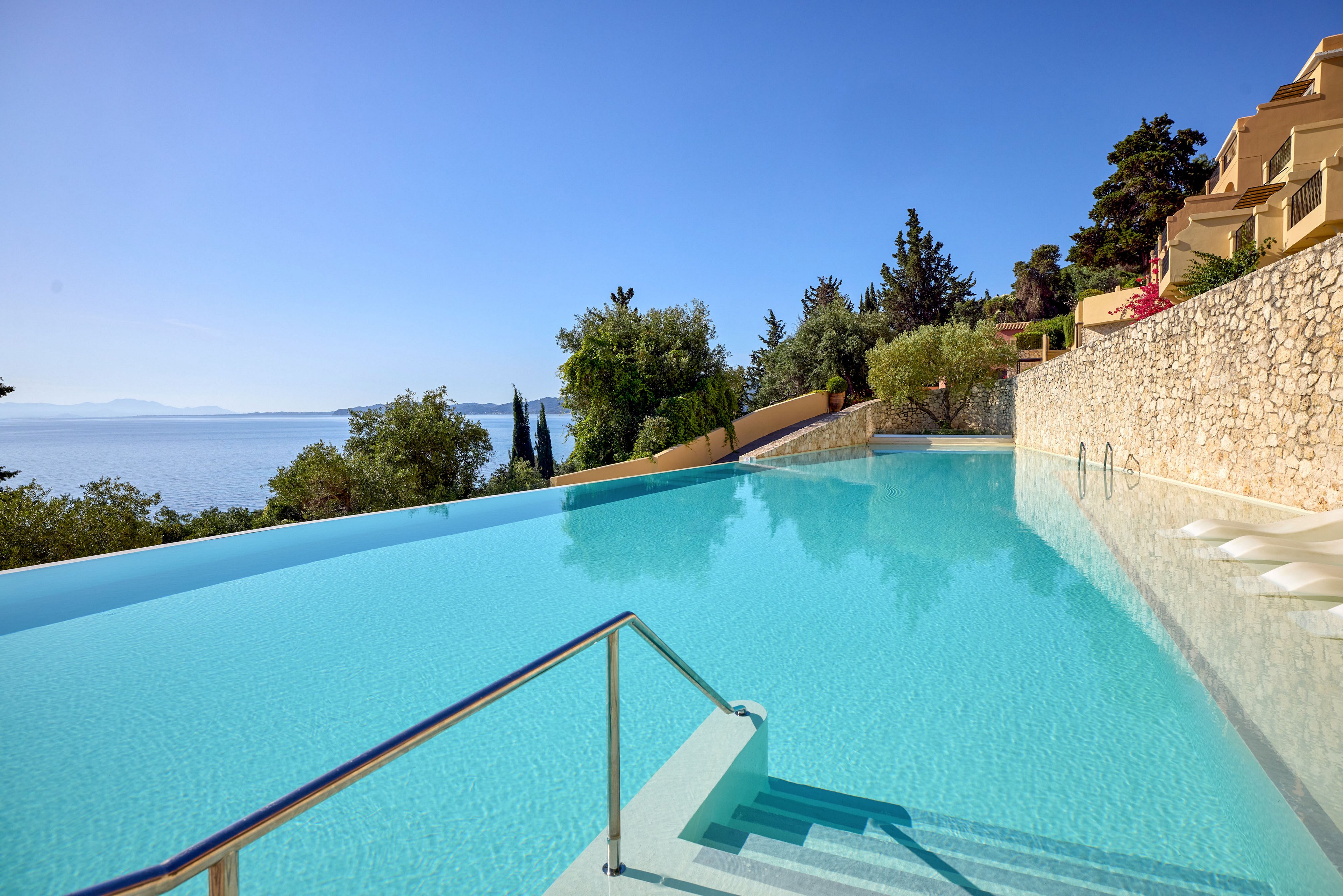 A tranquil infinity pool stretches toward a distant sea under a clear blue sky. Stone walls and terraced buildings frame the pool, creating a luxurious Mediterranean-style setting. The calm water reflects the sky, enhancing the sense of openness and relaxation. Lush greenery and coastal cliffs in the background complete the peaceful vacation atmosphere.