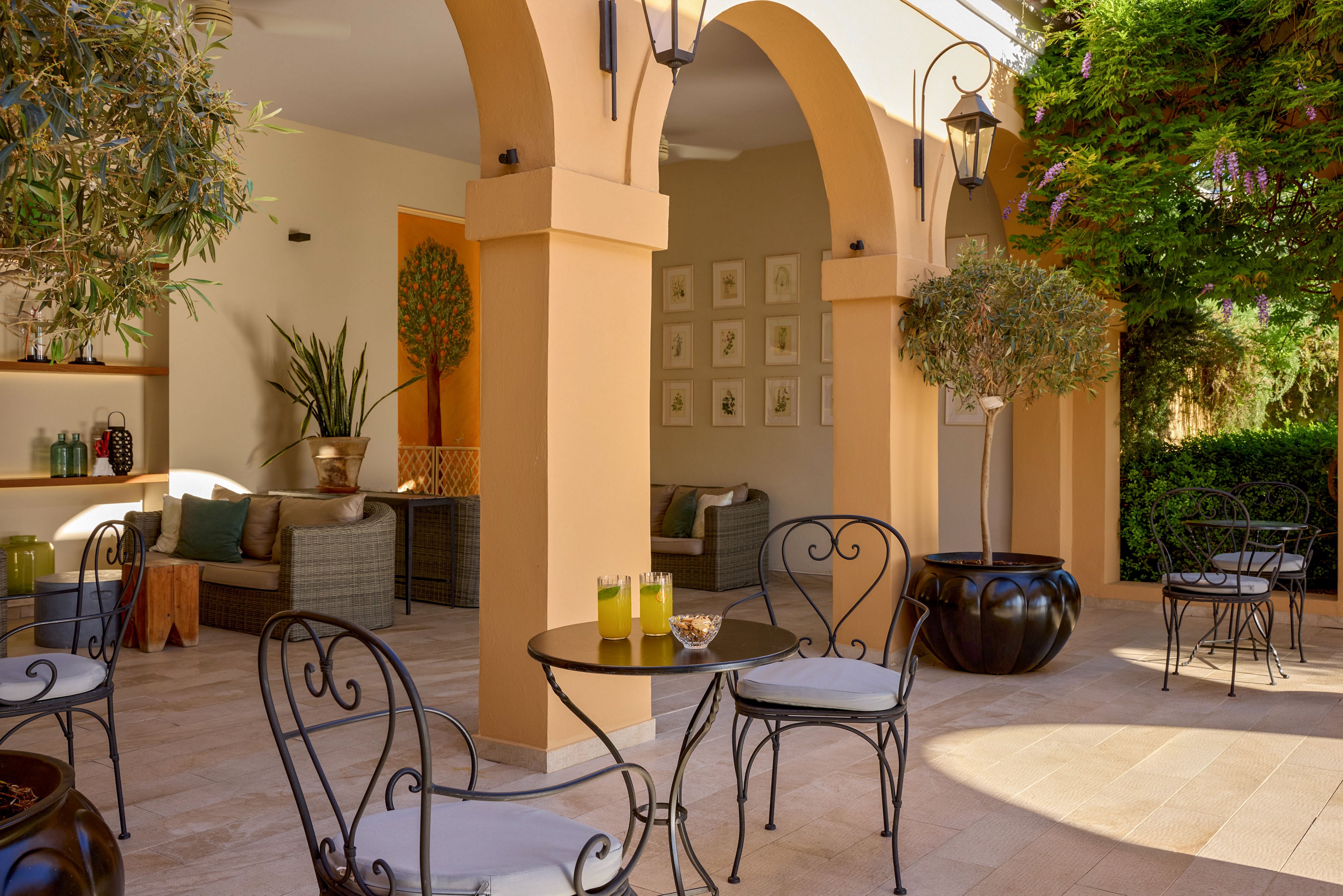 An inviting outdoor patio features wrought iron tables and chairs beneath warm, beige arches. Two glasses of orange juice and a glass of water sit on a small round table, suggesting a relaxed refreshment break. Comfortable wicker sofas with cushions are arranged in the shaded alcove, accented by potted plants and framed wall art. Large planters with small trees and lush greenery create a tranquil, Mediterranean-inspired atmosphere.