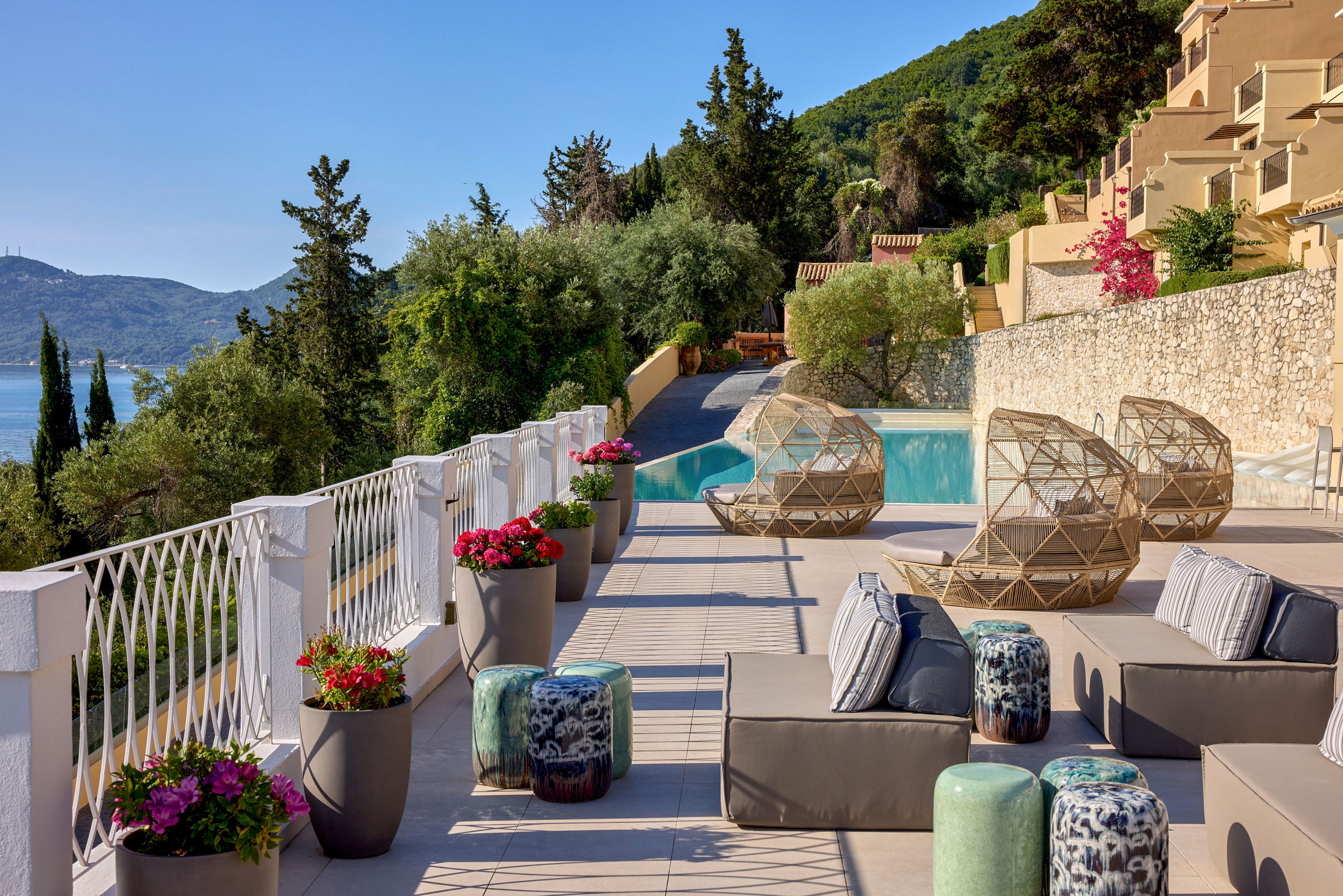 A stylish outdoor terrace overlooks a serene coastal landscape and forested hills. Contemporary lounge furniture and sculptural chairs are arranged beside a clear blue swimming pool. Colorful potted flowers line the white railing, adding vibrant accents to the neutral seating. Sunlit stone buildings rise along the hillside, creating an elegant Mediterranean resort atmosphere.