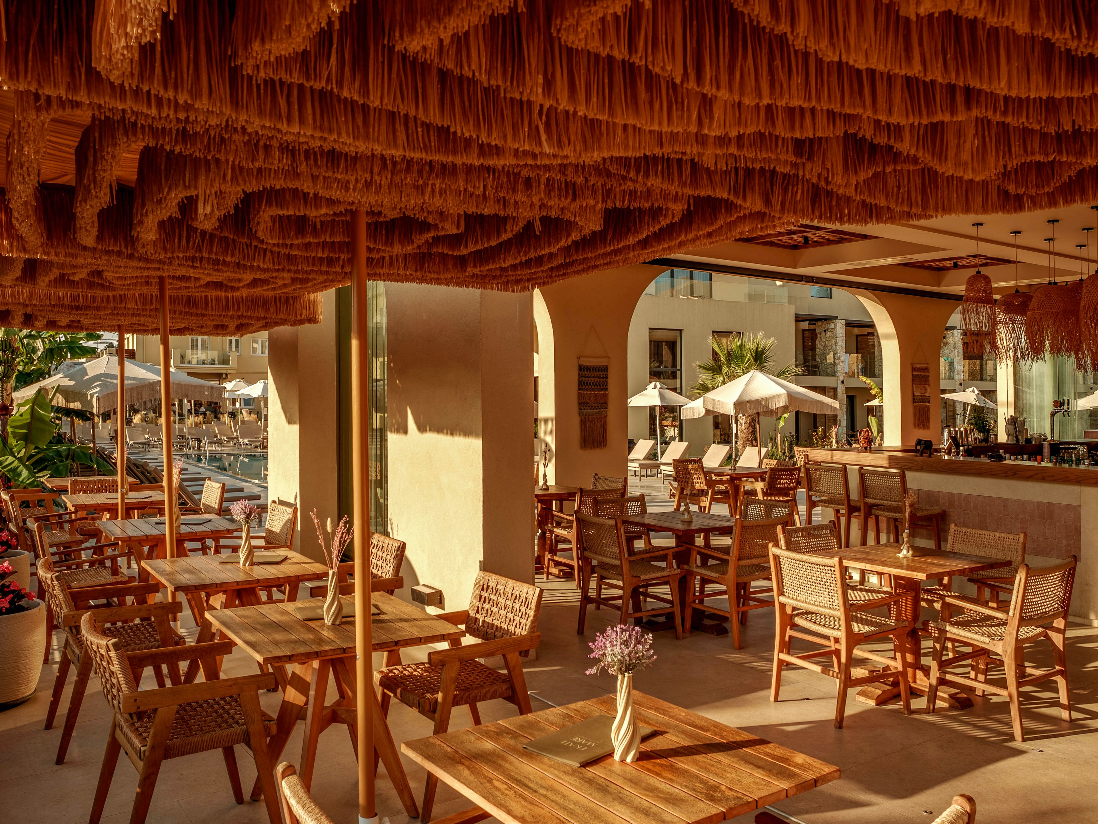 An open-air resort bar features neatly arranged wooden tables and woven chairs under a dramatic textured ceiling. Warm sunlight filters through the terrace, casting soft shadows across the floor and furniture. In the background, guests relax near a pool area with umbrellas and lounge chairs. The overall scene feels inviting and tranquil, ideal for outdoor dining or drinks in a vacation setting.