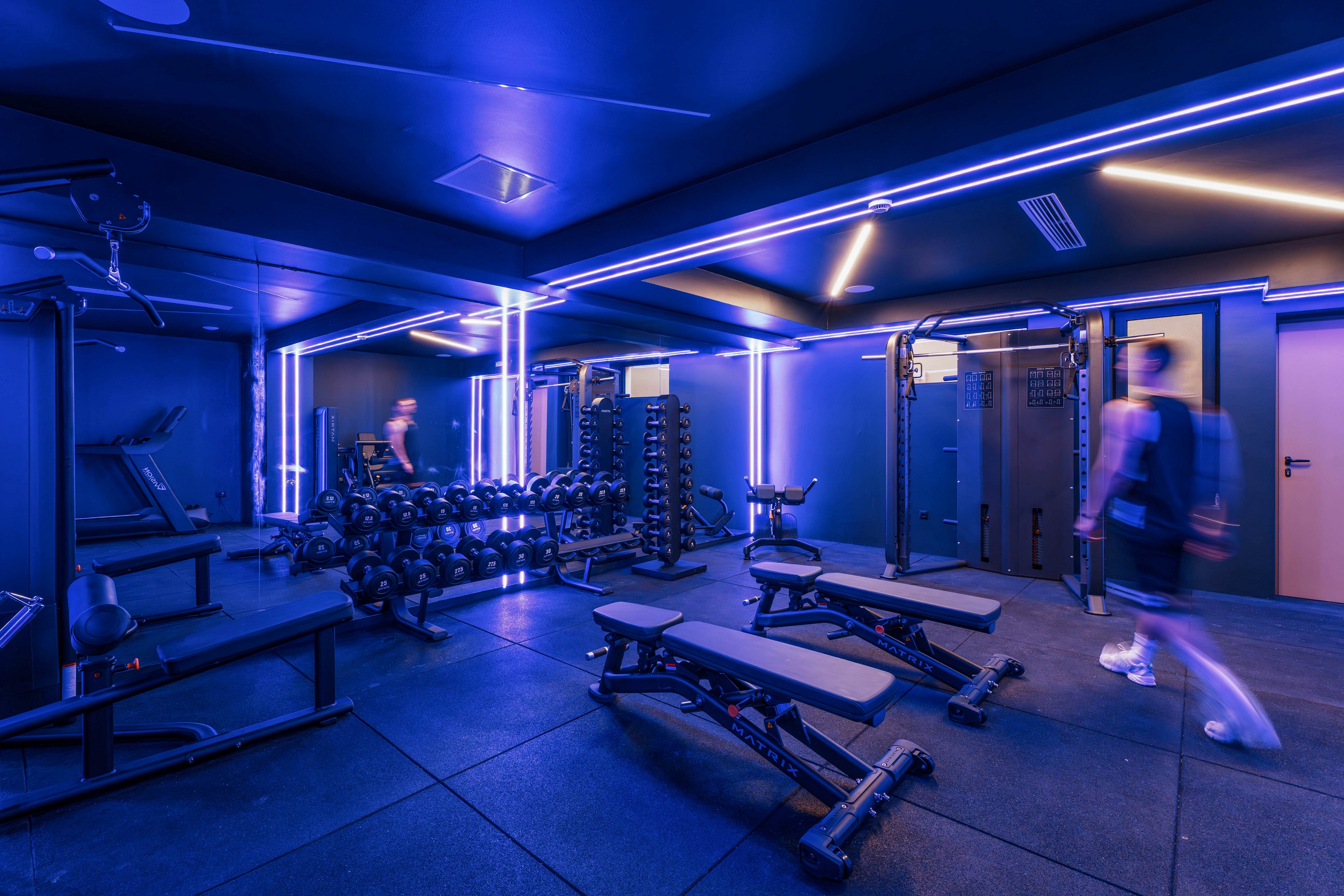 A contemporary gym interior is illuminated with vibrant blue neon strip lights along the ceiling and walls. Weight benches, dumbbells, and strength machines are neatly arranged on rubber flooring. Two blurred figures suggest people moving through the space, adding a sense of activity. The overall scene feels futuristic and energetic, emphasizing fitness in a stylish, modern environment.