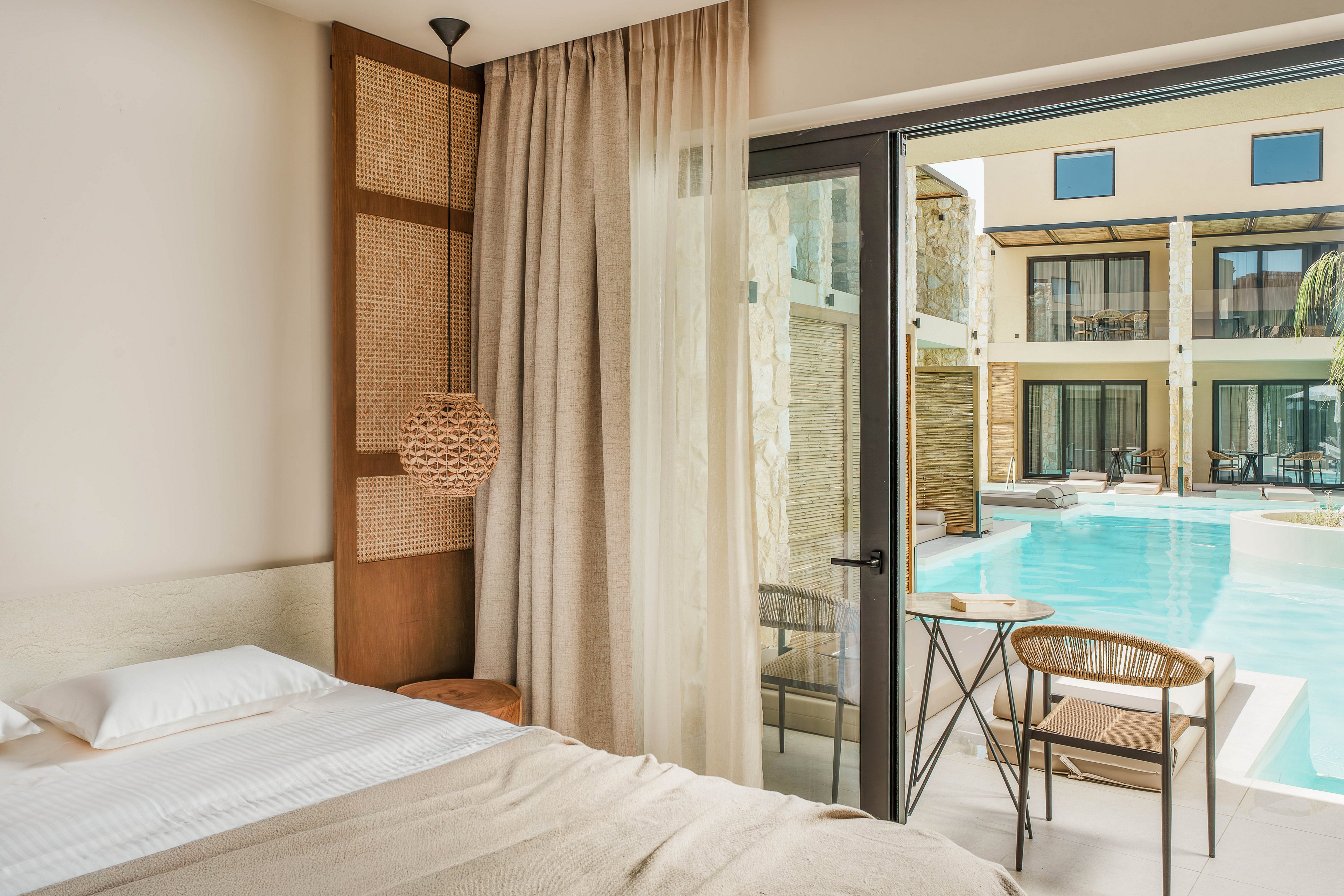 A contemporary hotel room features a neatly made bed with neutral linens and a woven accent headboard. Floor-to-ceiling curtains frame a large glass door that opens directly onto a poolside terrace. Outside, a small table and chairs sit beside a bright blue swimming pool, with neighboring rooms and balconies visible across the water. The overall scene feels calm, airy, and inviting, emphasizing resort-style relaxation.
