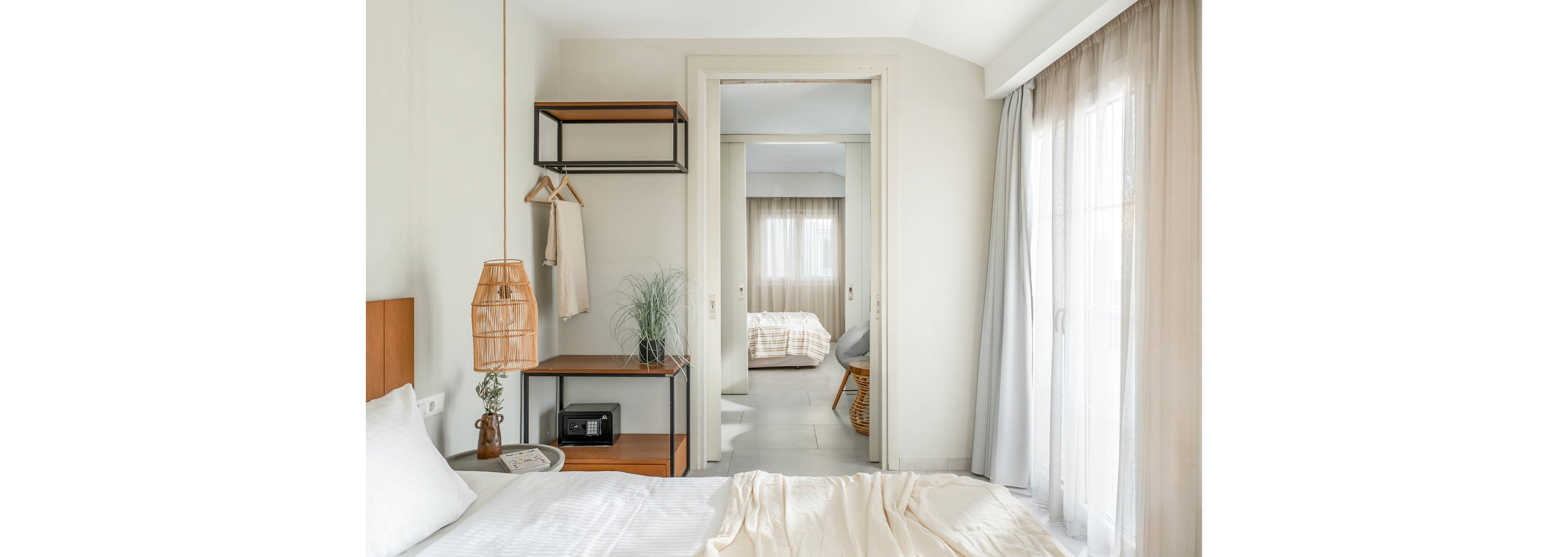 A bright minimalist bedroom features a neatly made bed with light bedding and soft neutral tones. An open shelving unit with a hanging rail, wicker lamp, potted plant, and small safe stands beside the bed. Sheer curtains filter daylight through large windows, creating an airy atmosphere. Through an open doorway, a second bedroom with similar decor and a wicker chair is visible, emphasizing a cohesive, modern interior design.