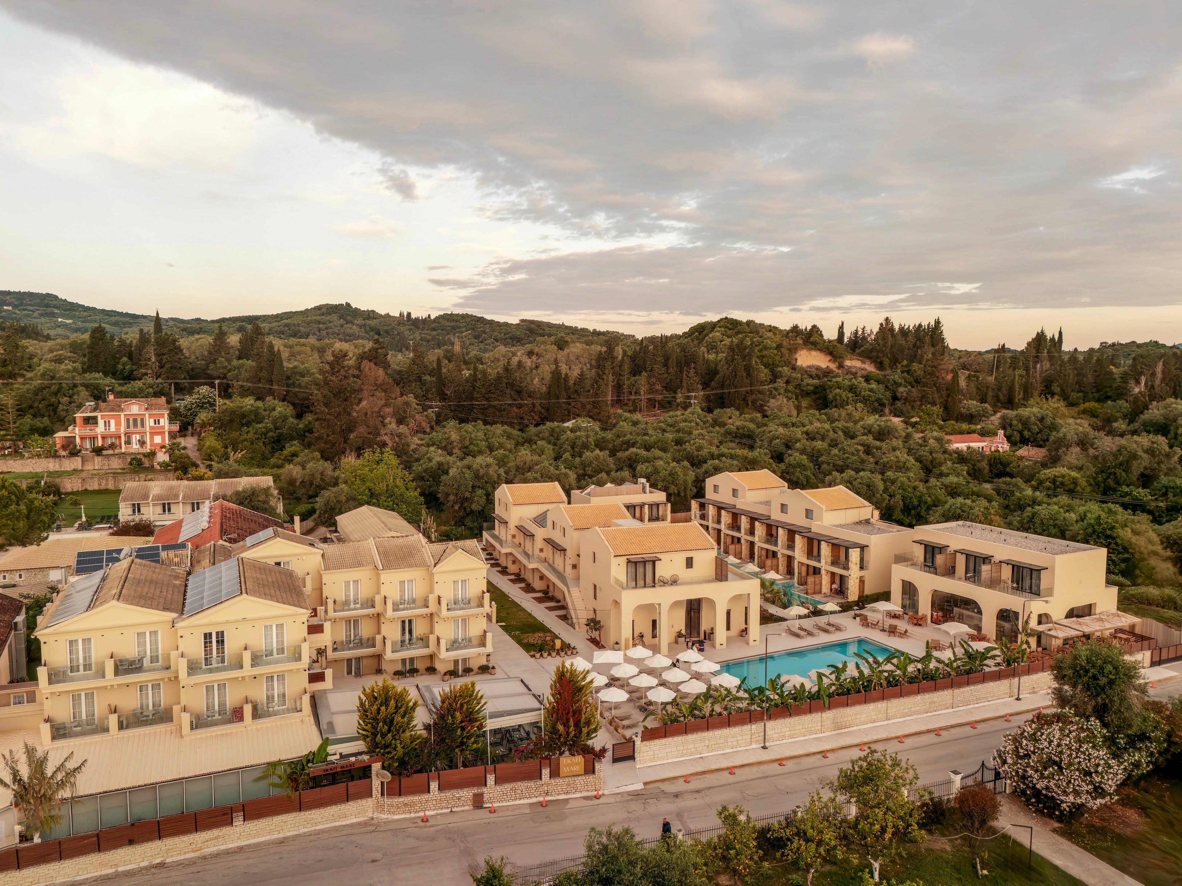 An aerial view shows a modern resort complex with multiple beige buildings and balconies arranged around a central swimming pool. The property is bordered by a road in the foreground and lush greenery and trees in the background. Poolside loungers and umbrellas line the water, suggesting a relaxing vacation setting. Soft evening or morning light and a cloudy sky create a calm, warm atmosphere.