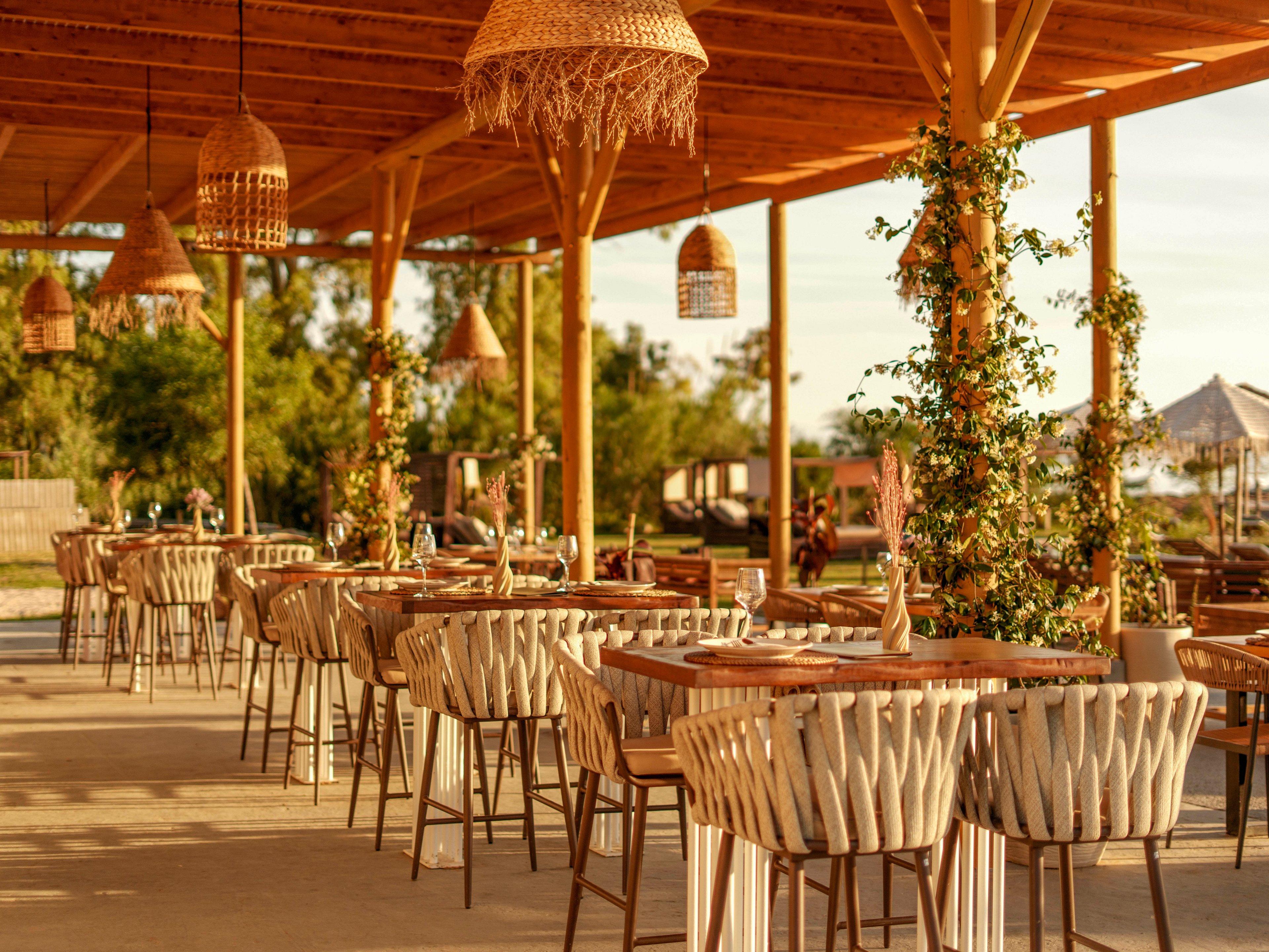An open-air restaurant terrace features rows of wooden tables and woven rope chairs under a wooden pergola. Warm sunlight creates a golden glow on the seating area and hanging wicker lamps. Green vines climb the pergola posts, adding a natural touch to the relaxed setting. In the background, trees and beach umbrellas suggest a coastal or resort location.