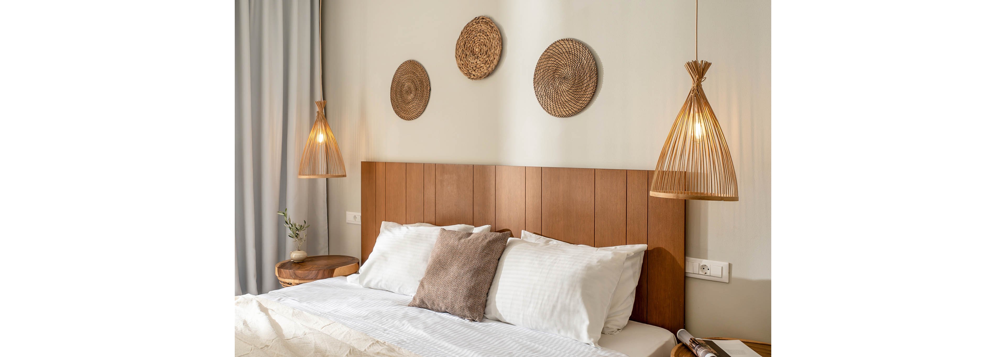 A neatly made bed with white linens and a brown accent pillow sits against a wooden headboard. The bedroom features warm hanging pendant lights with woven shades on either side of the bed. Round wicker wall hangings add texture and a natural touch above the headboard. Soft neutral tones and simple furnishings create a calm, minimalist atmosphere.