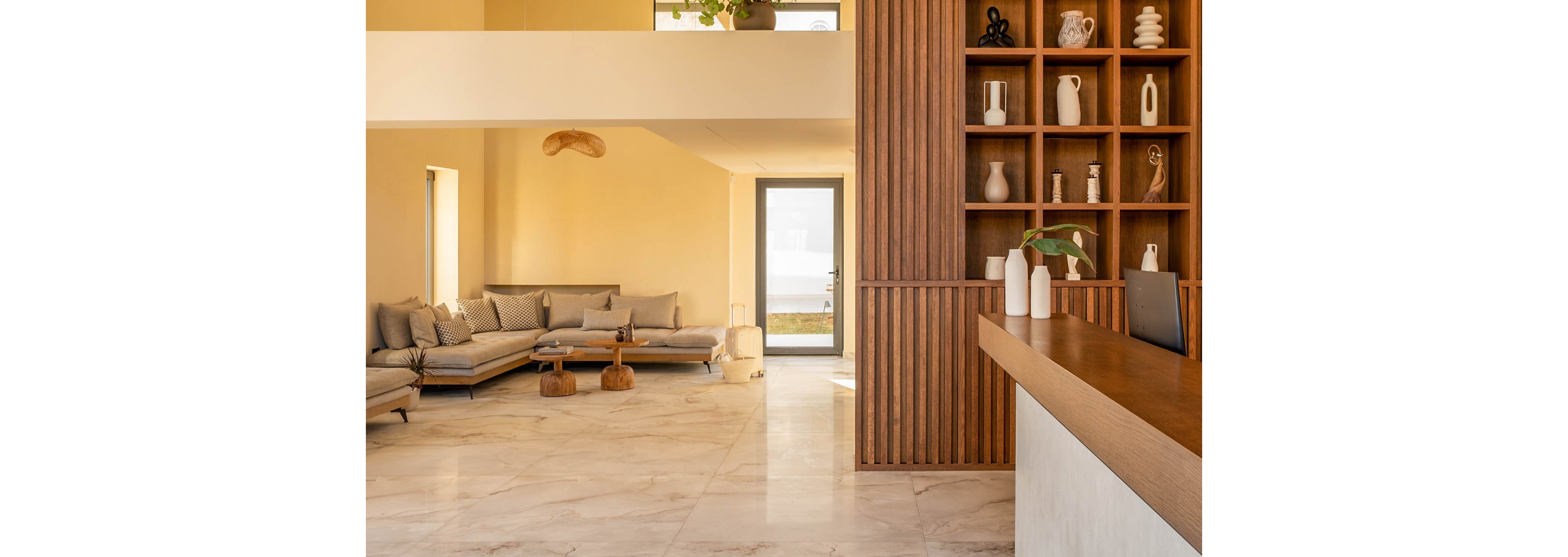 A modern, minimalist lobby features a wooden reception desk and built-in shelving filled with decorative vases and objects. To the left, a low-profile sectional sofa with neutral cushions creates a cozy seating area. Warm beige walls and large marble-look floor tiles enhance the airy, sunlit atmosphere. A glass door at the back reveals an outdoor view, adding depth and natural light to the space.