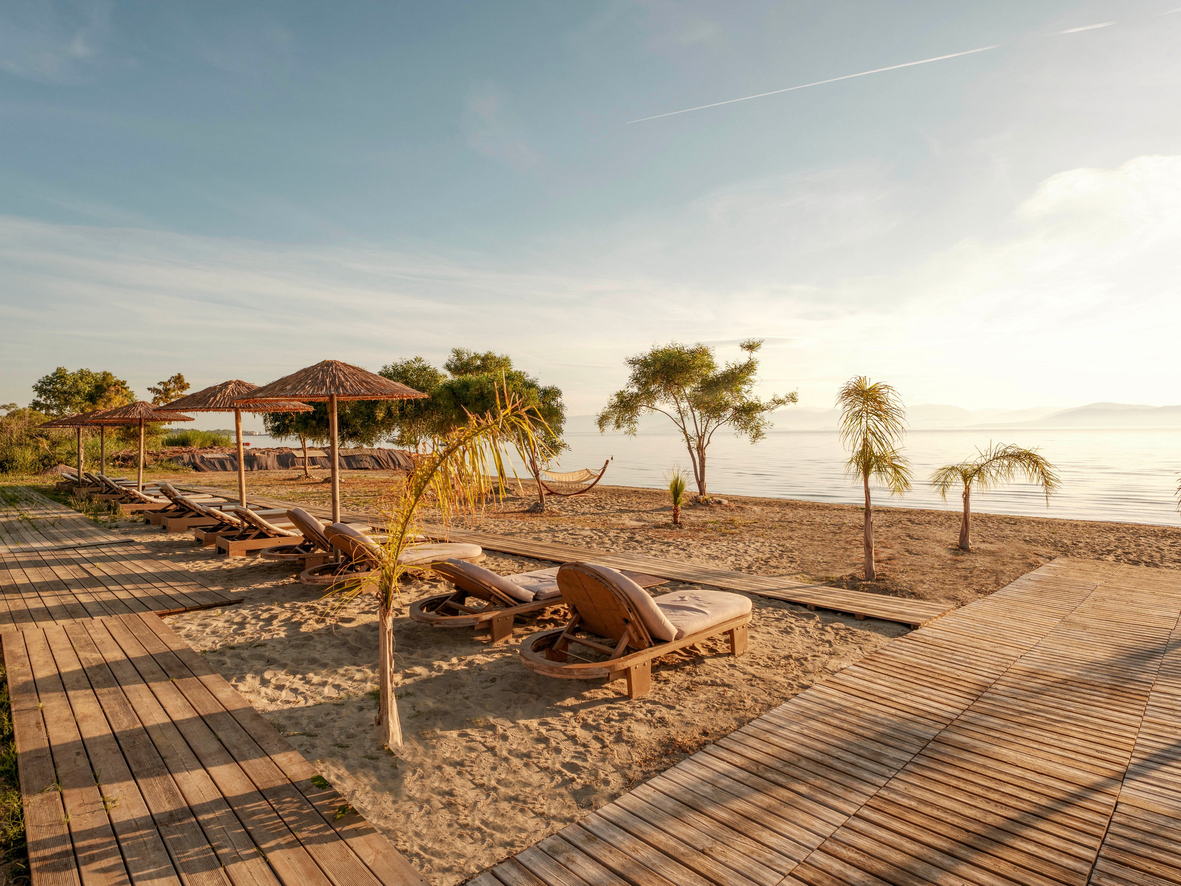 A tranquil sandy beach features several wooden sun loungers arranged along a boardwalk. Straw parasols and small palm trees provide shade near the shoreline. Calm sea water stretches into the distance under a soft, golden sunrise or sunset sky. The scene feels peaceful and inviting, with no people present and a hammock visible between trees.