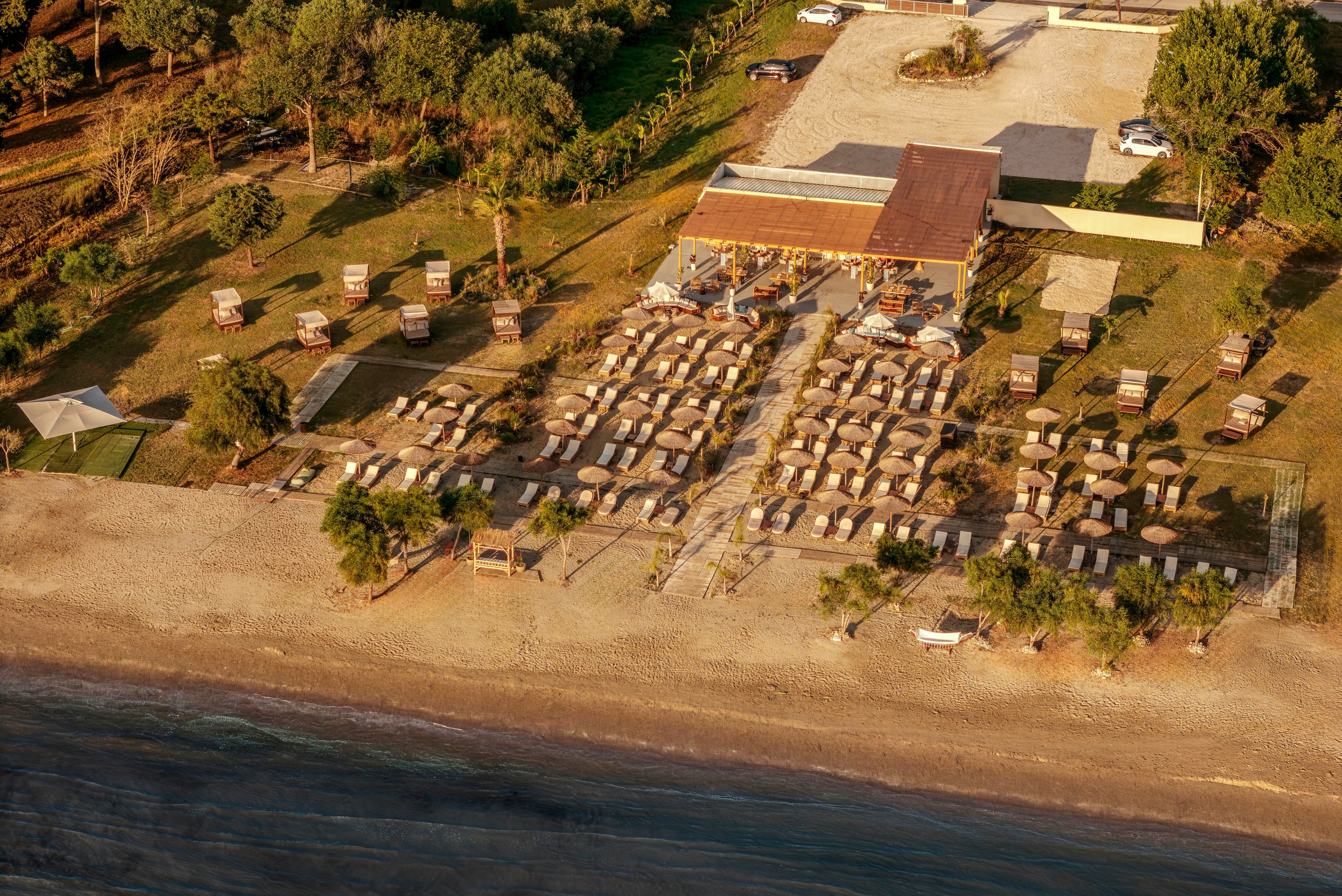An aerial view shows a neatly organized beachfront area with rows of sun loungers and umbrellas. A wooden beach bar or pavilion sits behind the loungers, surrounded by trees and greenery. The sandy shore stretches along the bottom of the image, meeting calm water. Warm sunlight casts long shadows, creating a relaxed, late-afternoon atmosphere.
