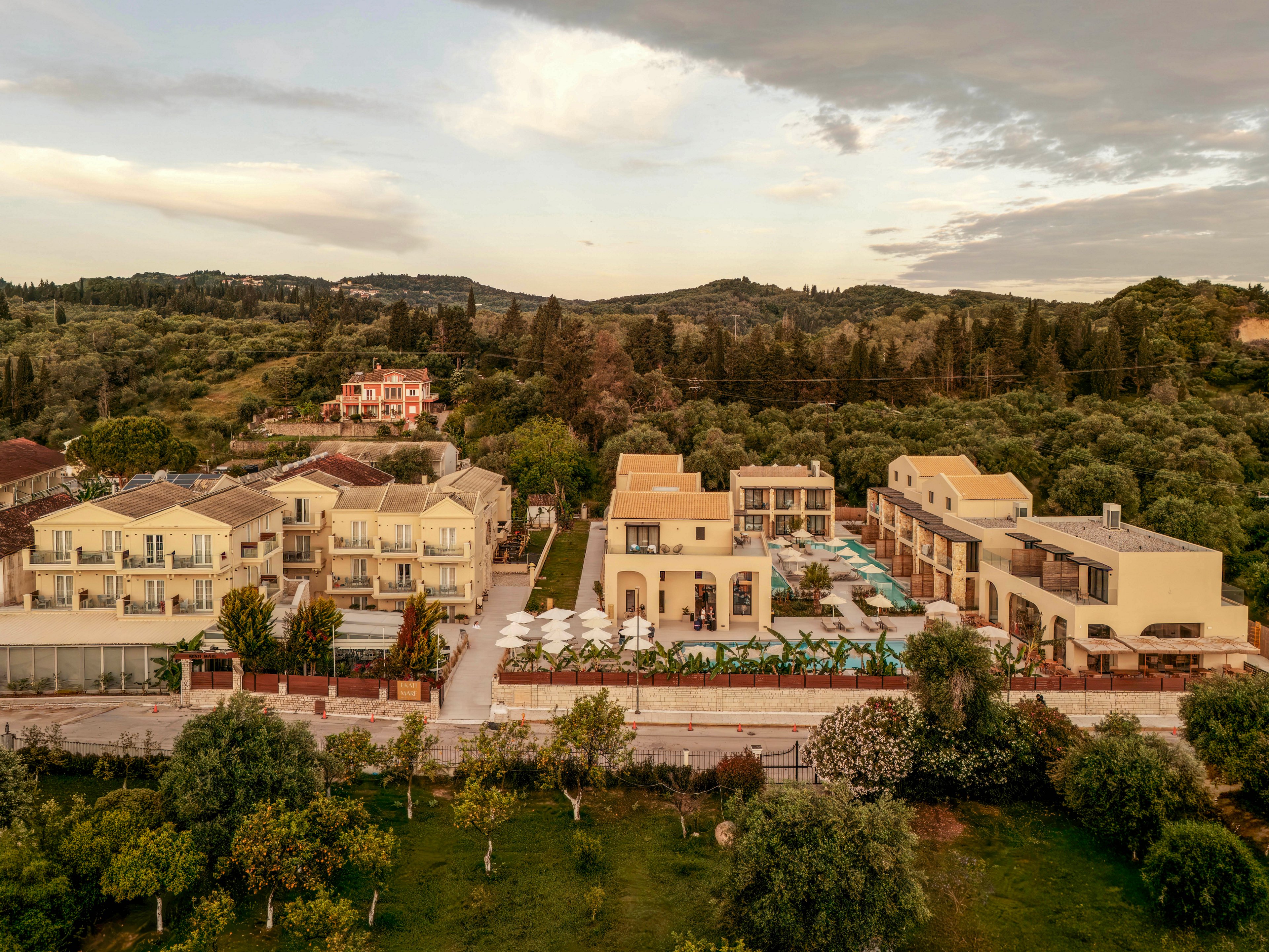 An aerial view shows a modern resort complex with multiple beige buildings arranged along a main road. The property features several swimming pools, sun loungers, and umbrellas in a landscaped courtyard. Surrounding the resort are lush green fields, olive trees, and forested hills stretching into the distance. Warm evening light and a soft sky create a tranquil vacation atmosphere.