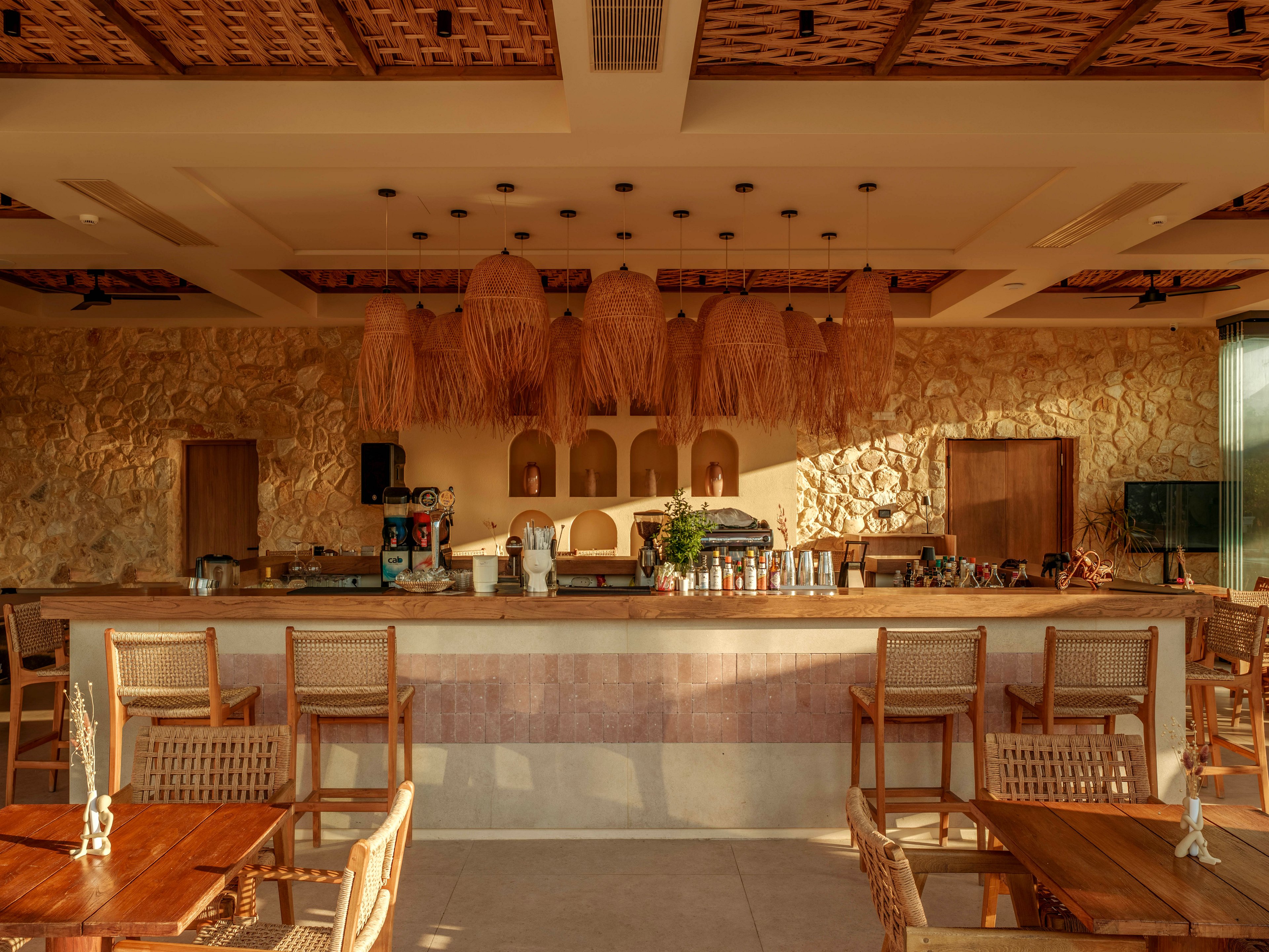 A cozy resort bar interior features a long counter framed by stone walls and wooden accents. Woven pendant lights hang from the ceiling, casting a warm golden glow across the space. Wooden tables and chairs with woven backs are arranged neatly in the foreground, facing the bar. Bottles, glassware, and bar equipment line the countertop, suggesting a relaxed vacation setting at sunset.