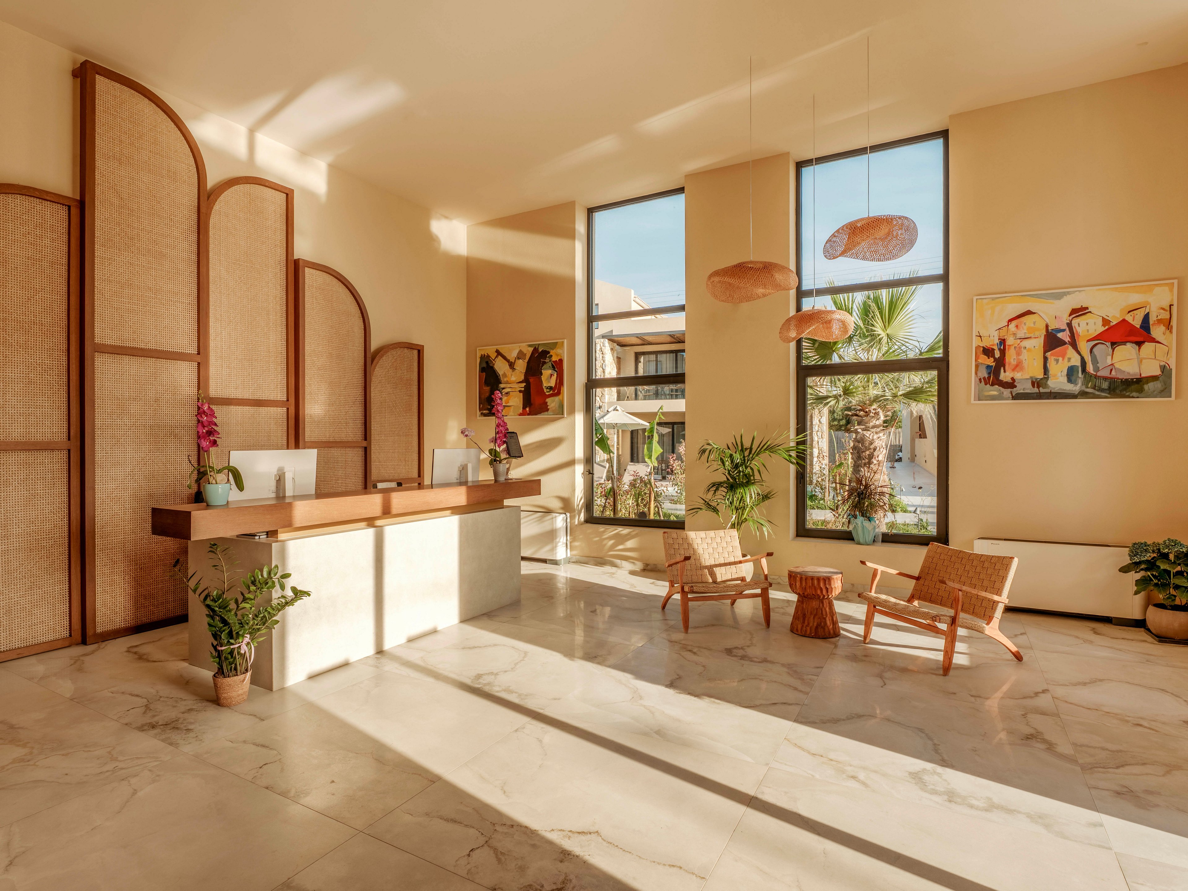A bright, modern hotel lobby features a minimalist reception desk with two computers and decorative orchids. Large floor-to-ceiling windows let in abundant sunlight and reveal an outdoor courtyard with palm trees. Woven chairs and a small table create a relaxed seating area on the polished marble floor. Warm neutral tones, natural wood accents, and contemporary artwork contribute to a calm, welcoming atmosphere.