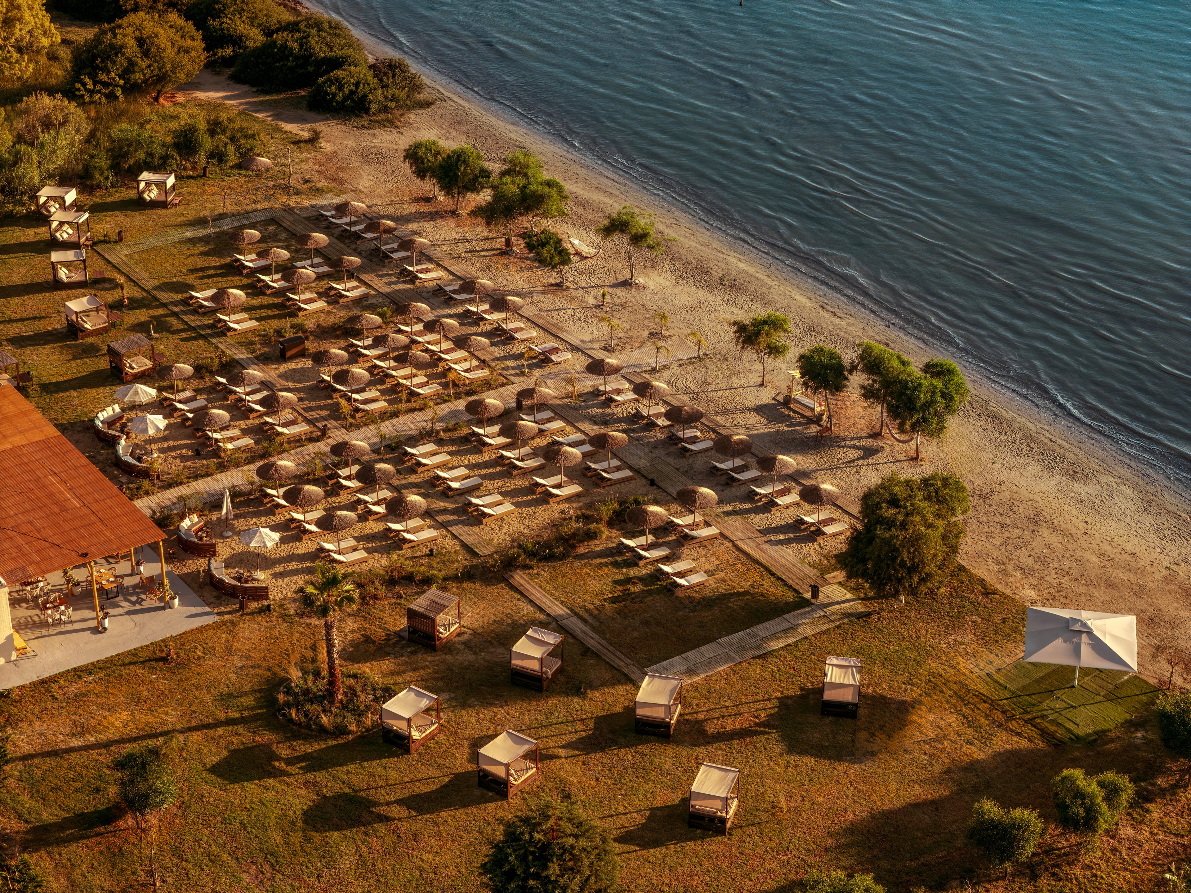 An aerial view shows a neatly arranged beachfront resort with rows of sun loungers and umbrellas facing the sea. The shoreline curves gently along calm blue water, with a narrow strip of sand separating the beach from the grassy resort grounds. Small cabanas and shaded seating areas are scattered across the lawn, framed by trees and landscaped greenery. Warm golden light suggests either sunrise or sunset, creating a peaceful and inviting vacation atmosphere.