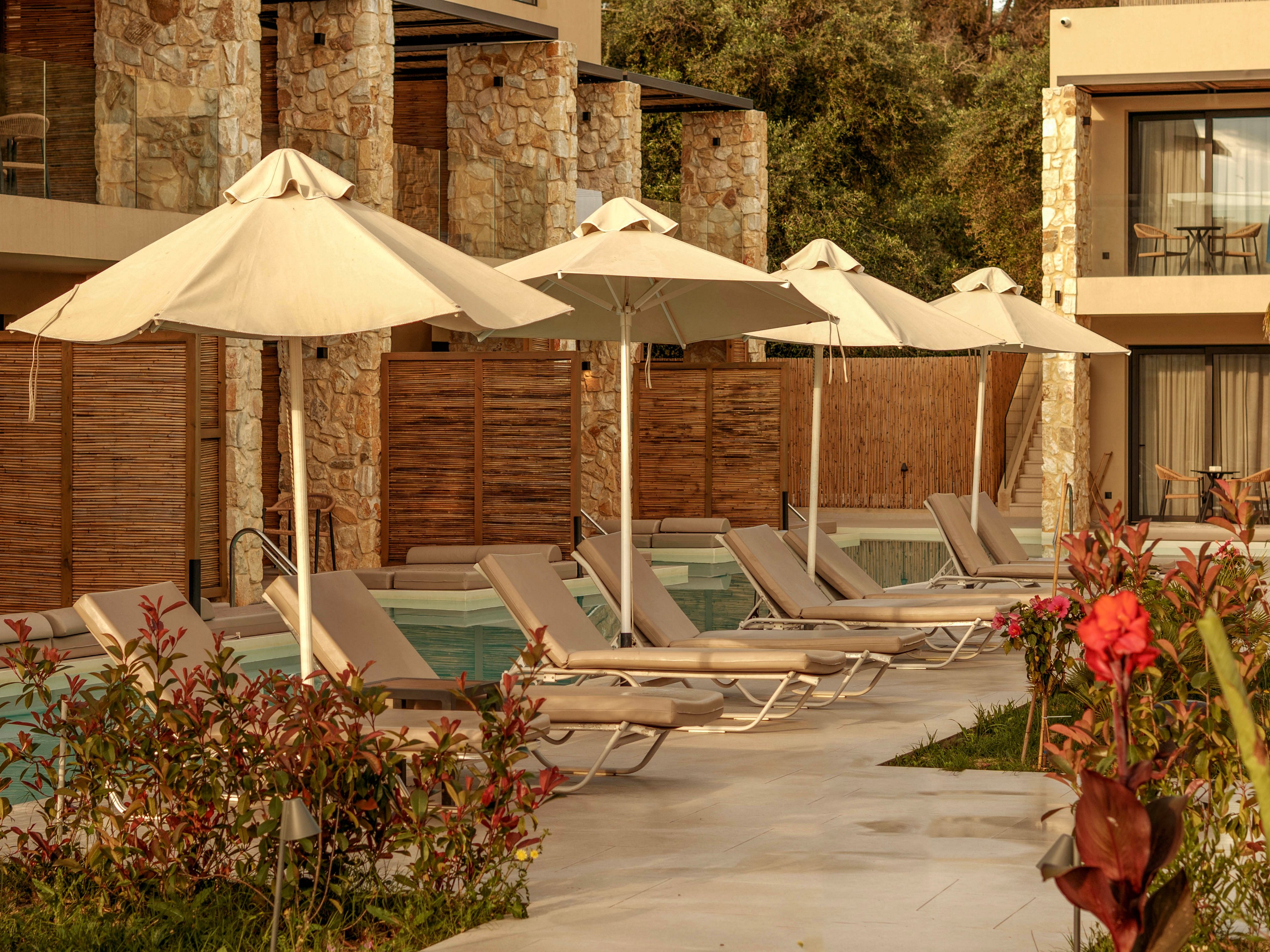 A row of beige sun loungers lines a narrow poolside area in front of a modern resort building. Large white umbrellas provide shade along the walkway, creating a relaxed vacation atmosphere. Stone and wood architectural details frame the balconies and rooms behind the pool. Lush green plants and red flowers add color and softness to the warm-toned scene.