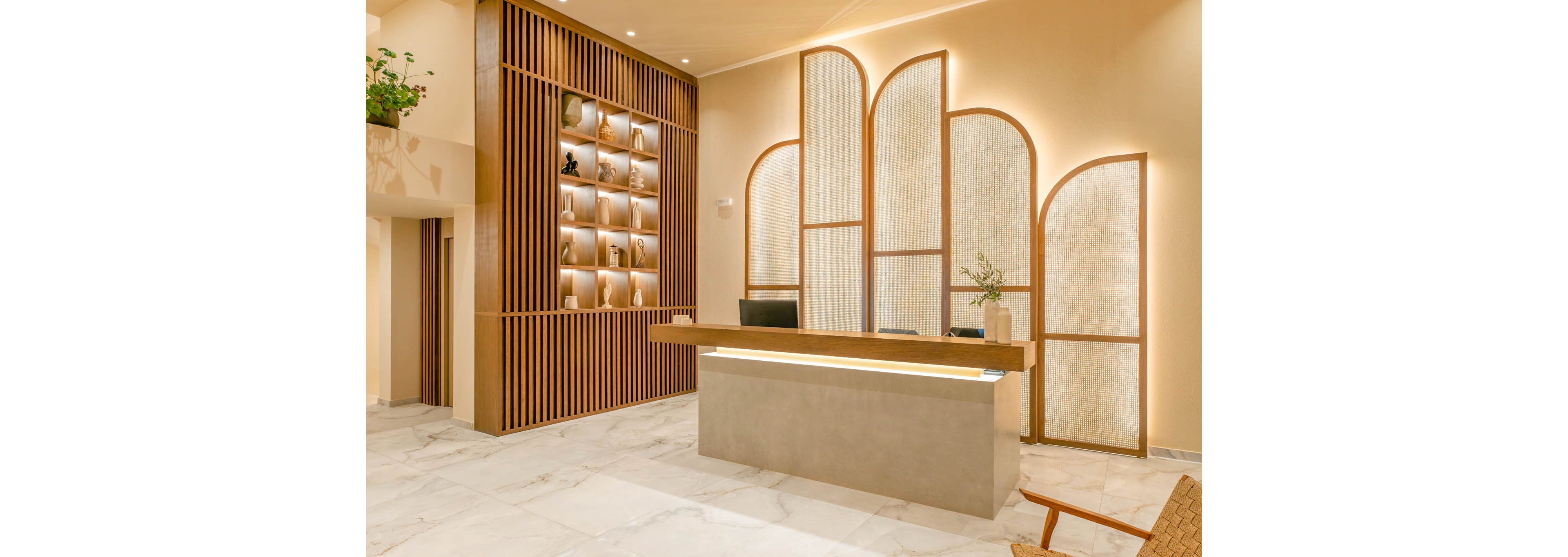 A contemporary reception desk stands in a bright, minimalist lobby. The space features warm beige walls, marble-style flooring, and a sleek counter with a computer monitor. A decorative wooden wall with illuminated niches displays small objects, while curved panel screens add texture behind the desk. Soft, indirect lighting and a few plants create a calm, welcoming atmosphere.