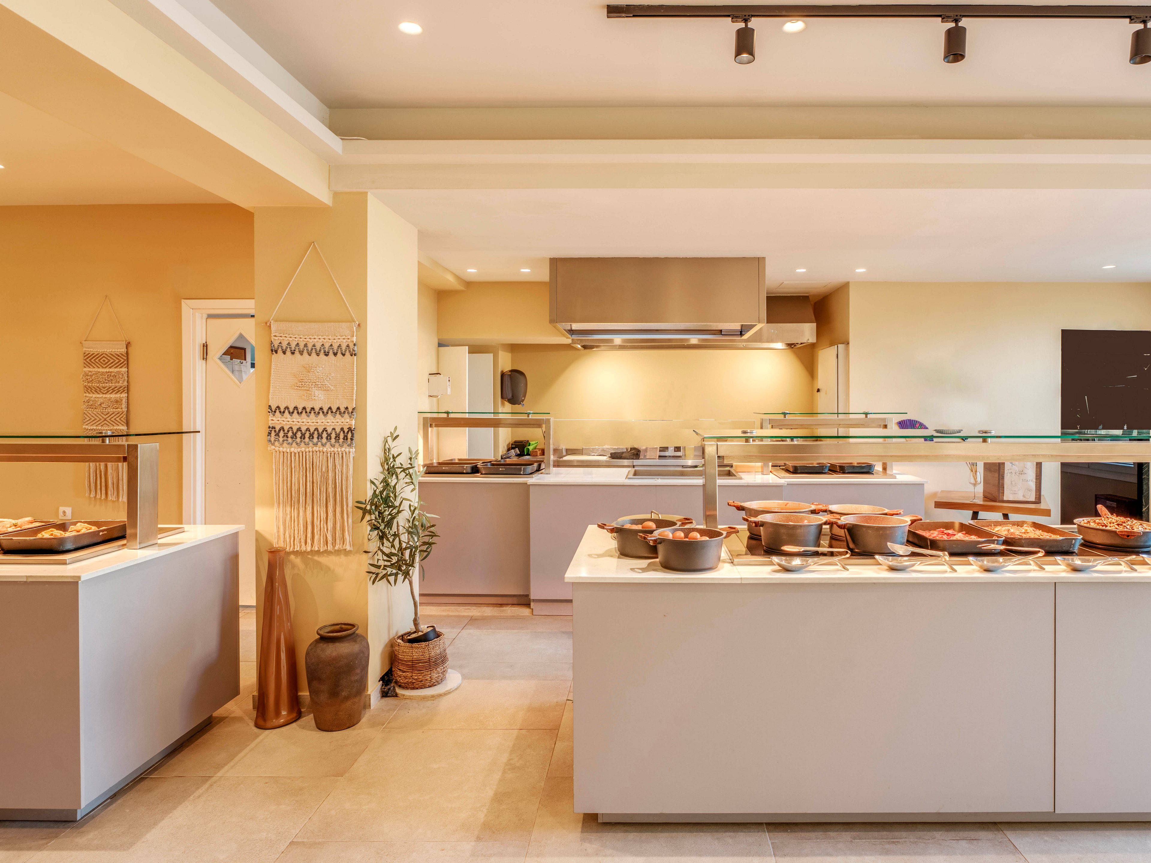 A contemporary self-service buffet counter is set up in a bright, warm-toned dining area. Various dishes in pots and trays are arranged neatly under glass sneeze guards. The background features a stainless steel range hood, minimalist cabinetry, and subtle decorative elements like woven wall art and ceramic vases. Soft lighting and neutral colors create a welcoming, relaxed atmosphere suitable for hotel or restaurant dining.
