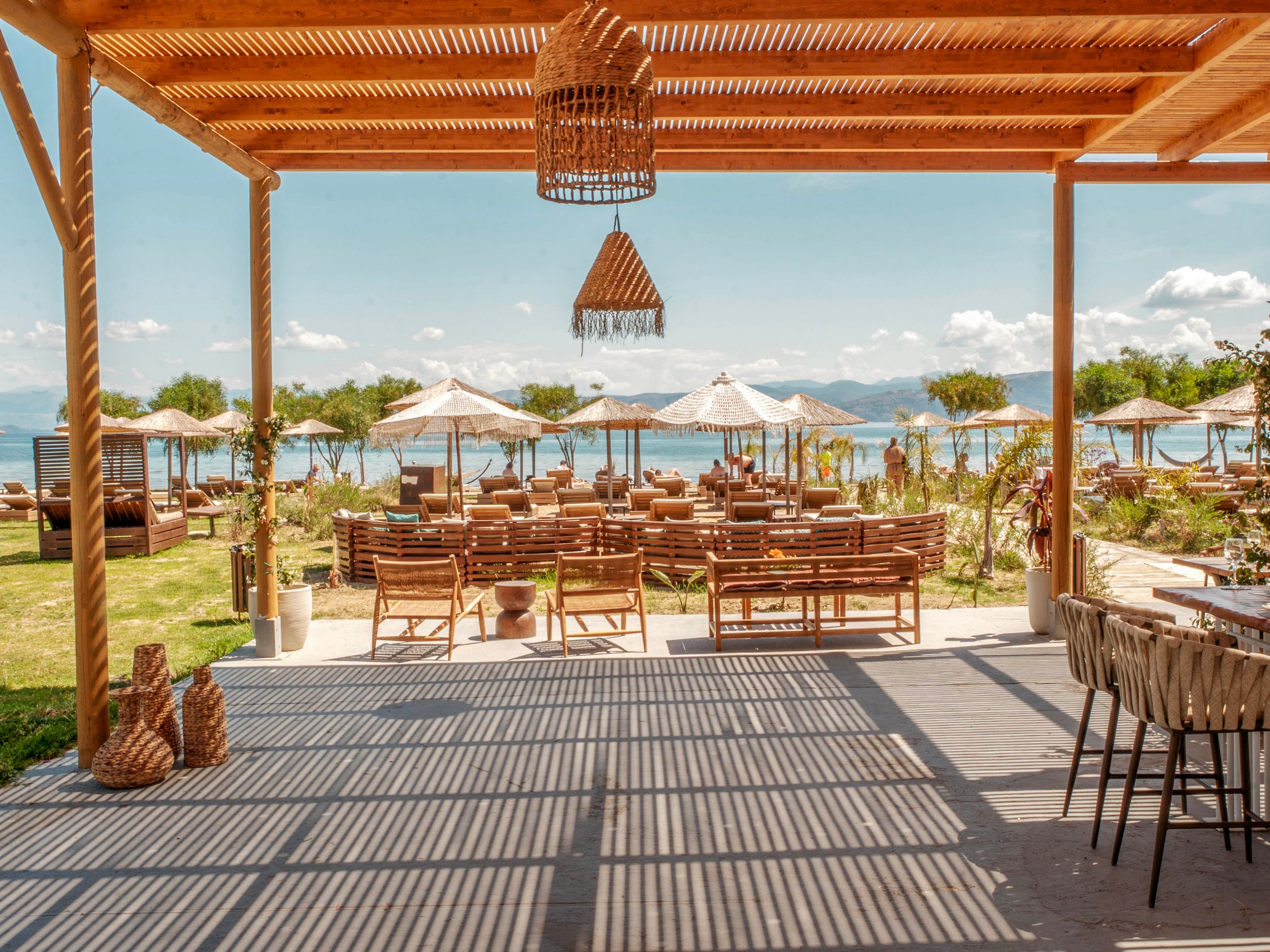 An open-air wooden terrace overlooks a sandy beach and calm sea. Rows of wooden tables and chairs are arranged under a pergola casting striped shadows on the floor. Straw umbrellas and sun loungers line the shoreline in the background. The scene feels relaxed and summery, with warm natural tones and clear blue sky.