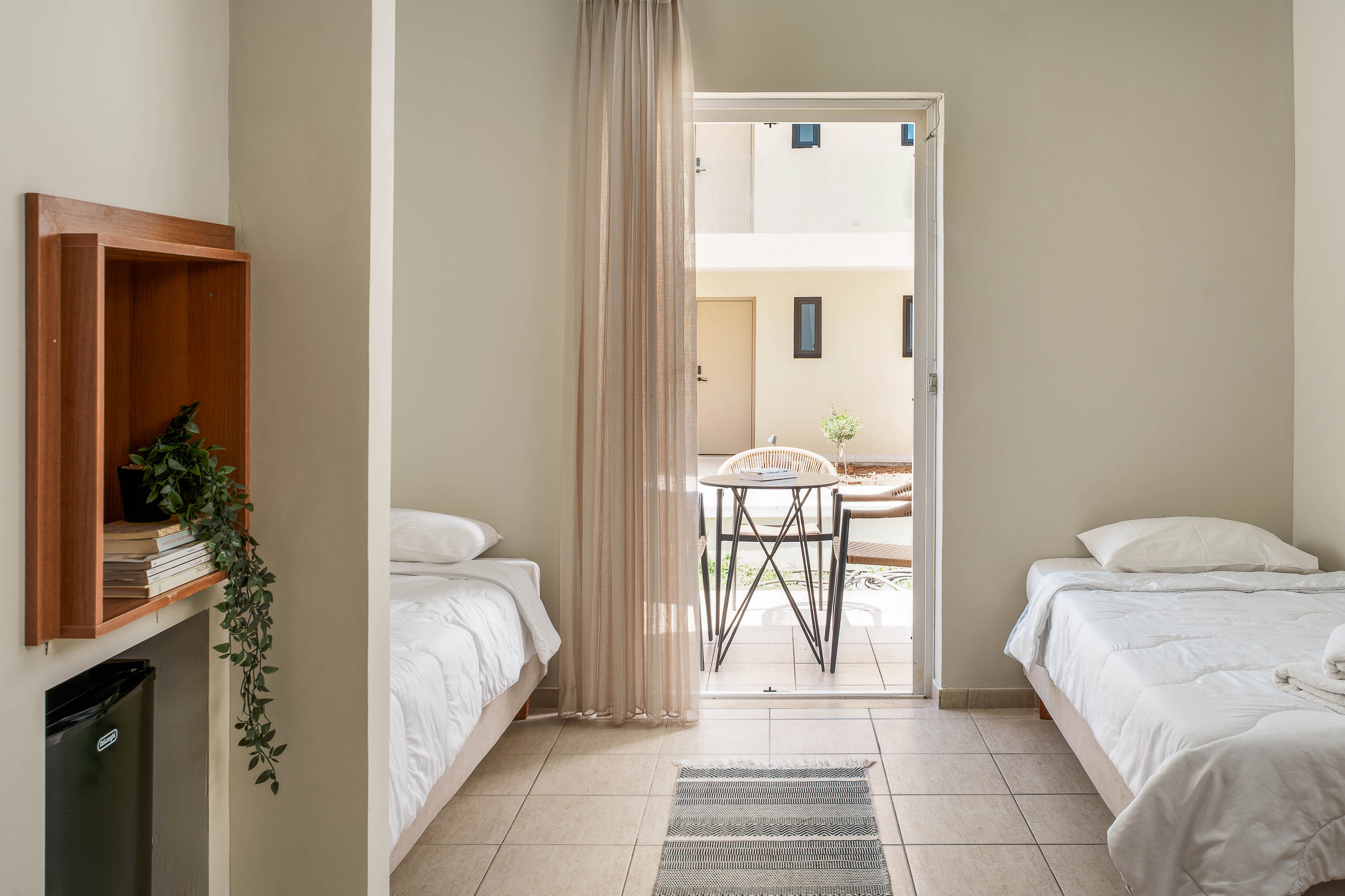 A simple twin bedroom features two single beds with white linens facing an open doorway. Sheer curtains frame the entrance to a small balcony with a round table and two chairs. Neutral walls and tiled flooring create a clean, minimalist atmosphere. A small shelf with books and a potted plant and a compact refrigerator add functional details to the cozy space.