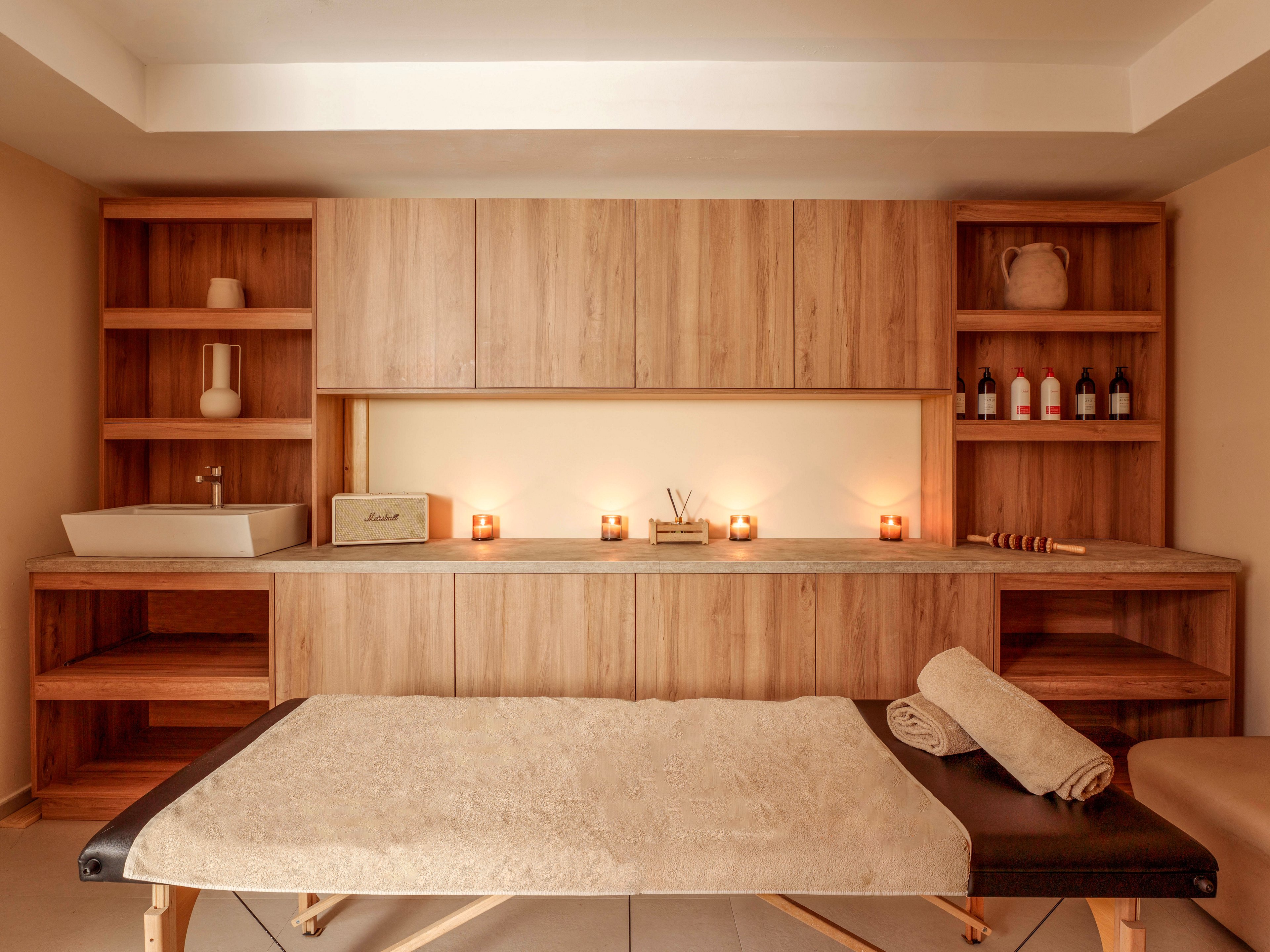 A cozy spa treatment room features a massage table in the foreground covered with a neutral-toned sheet and rolled towels. Built-in wooden cabinets and open shelving line the back wall, holding decorative vases, bottles, and spa accessories. Several lit candles create a soft, ambient glow along the countertop, enhancing the relaxing atmosphere. The overall palette is warm and earthy, emphasizing comfort and tranquility.