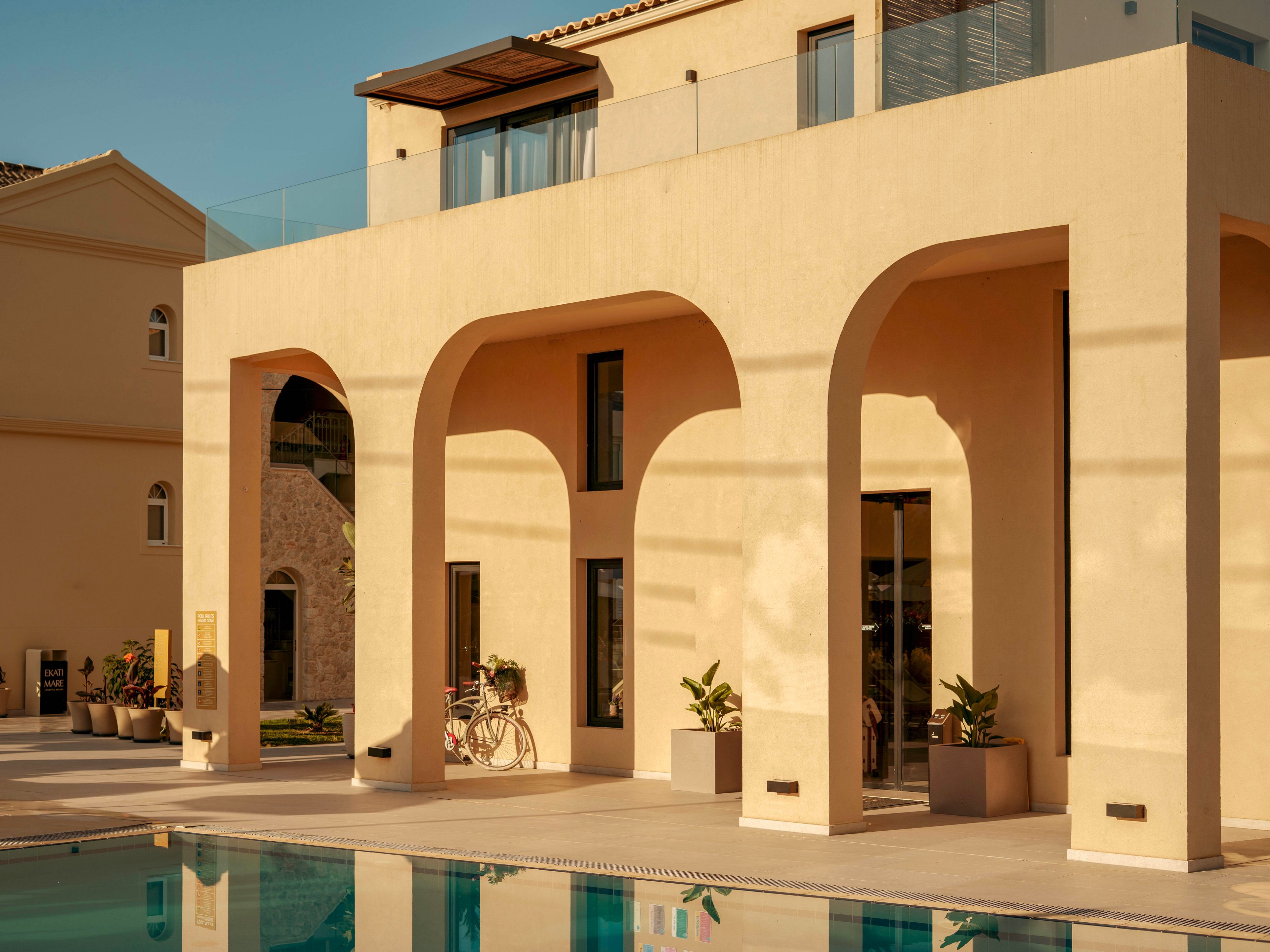 A contemporary villa with large arches stands beside a clear swimming pool in warm evening light. The architecture features smooth beige walls, tall windows, and a minimalist terrace. Potted plants and a bicycle add casual, resort-like details to the scene. Soft shadows and golden tones create a relaxed, luxurious holiday atmosphere.