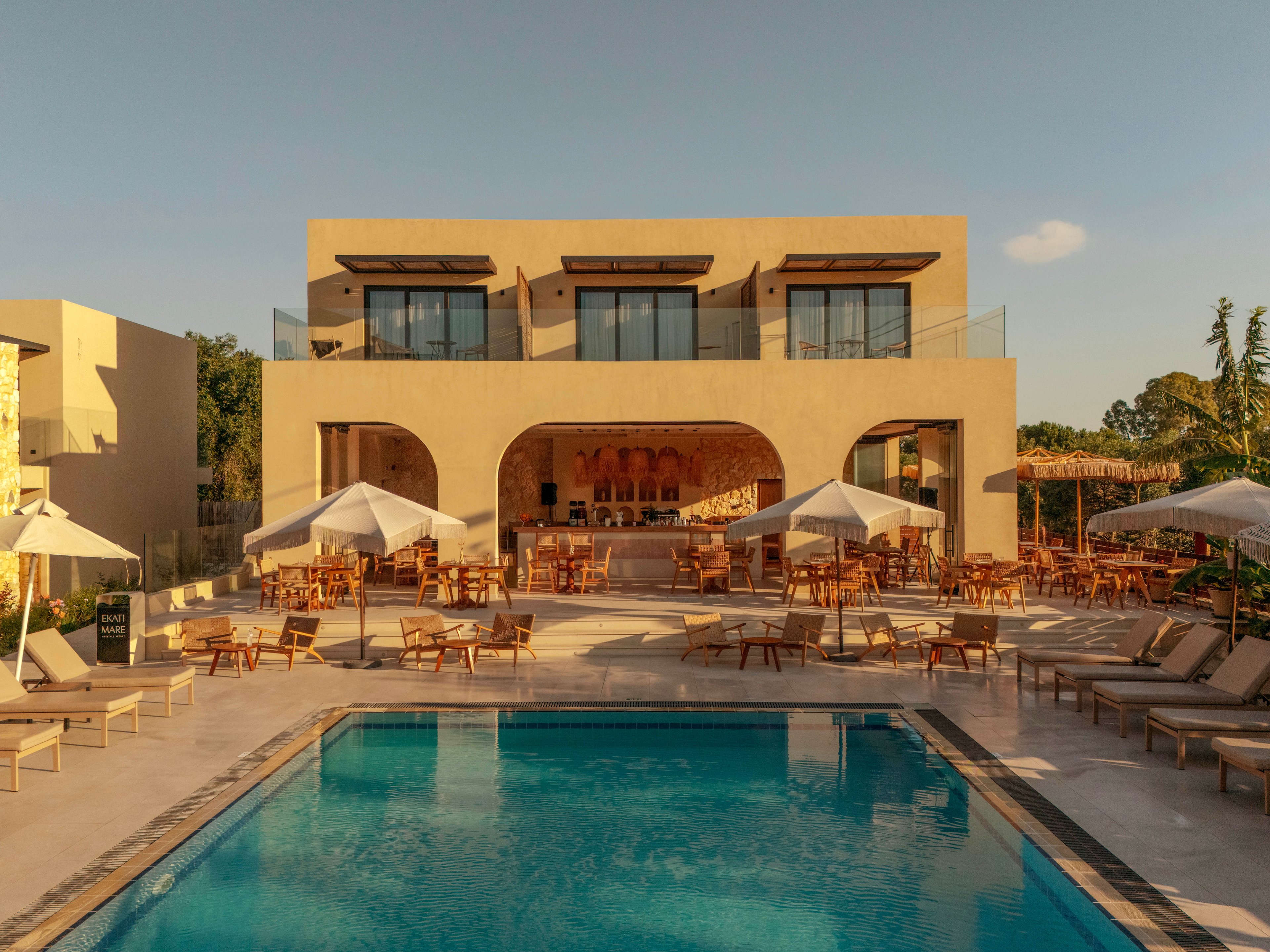 A contemporary two-story resort building overlooks a clear blue swimming pool. Wooden lounge chairs and white umbrellas are arranged neatly around the pool, creating an inviting outdoor seating area. Warm beige architecture and natural wood tones are highlighted by the golden hour sunlight. The scene suggests a relaxed vacation atmosphere in a stylish, modern hotel setting.