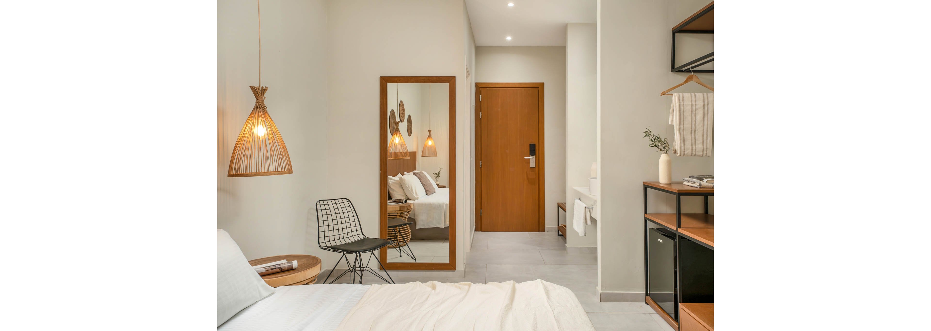 A contemporary hotel bedroom features a neatly made bed with light-colored linens and a wooden side table. A wireframe chair sits beside a large floor mirror reflecting the bed and wall decor. Warm wooden accents, including the door and mirror frame, contrast with the neutral walls and tiled floor. Soft lighting from a woven pendant lamp and recessed ceiling lights creates a calm, inviting atmosphere.