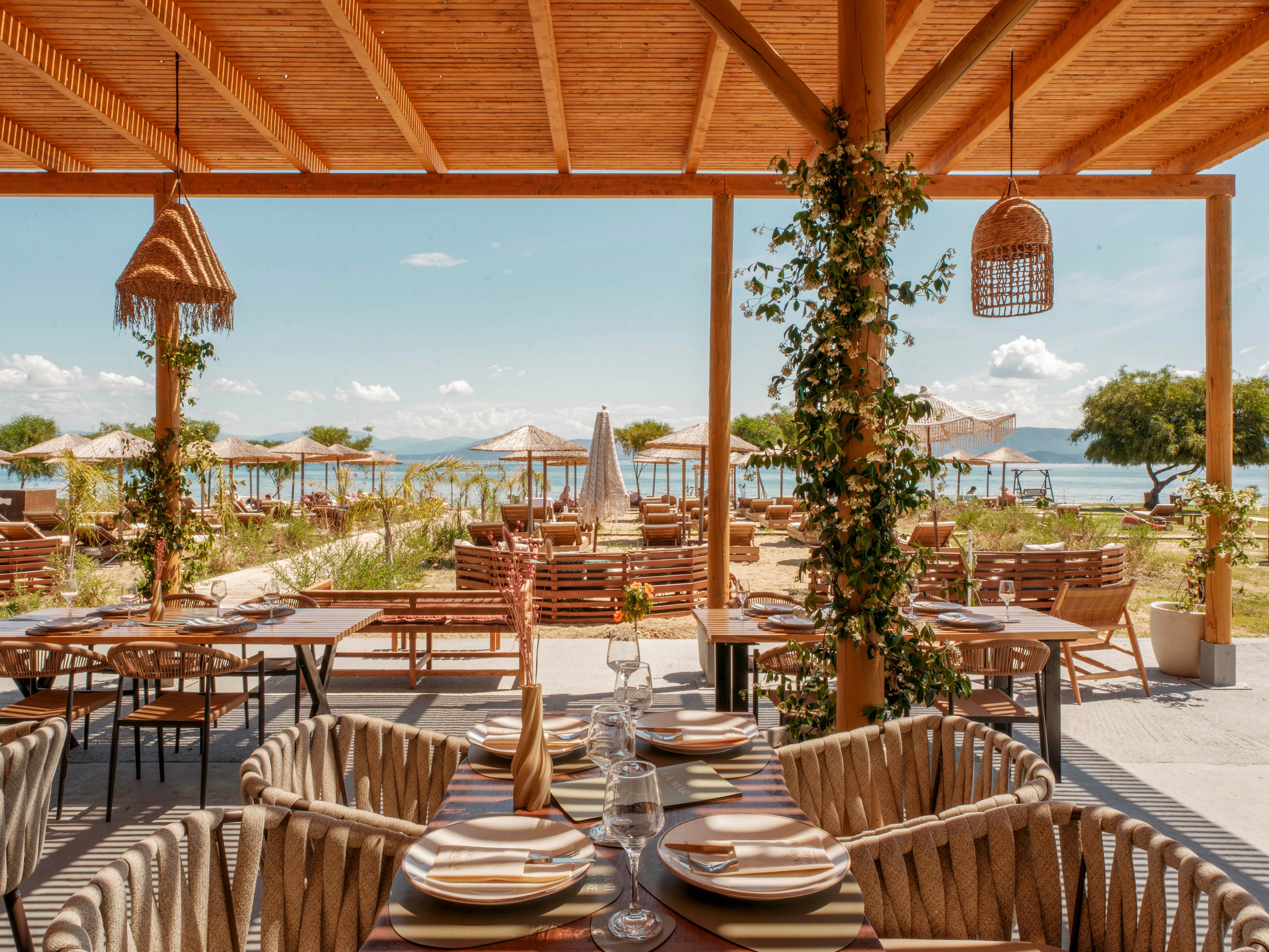 An open-air restaurant terrace overlooks a sandy beach and calm sea. Wooden pergolas, woven chairs, and neatly set tables create a relaxed coastal dining atmosphere. Sun loungers and umbrellas line the beach in the background under a bright blue sky. Green plants climb the wooden posts, adding a natural touch to the warm, earthy color palette.