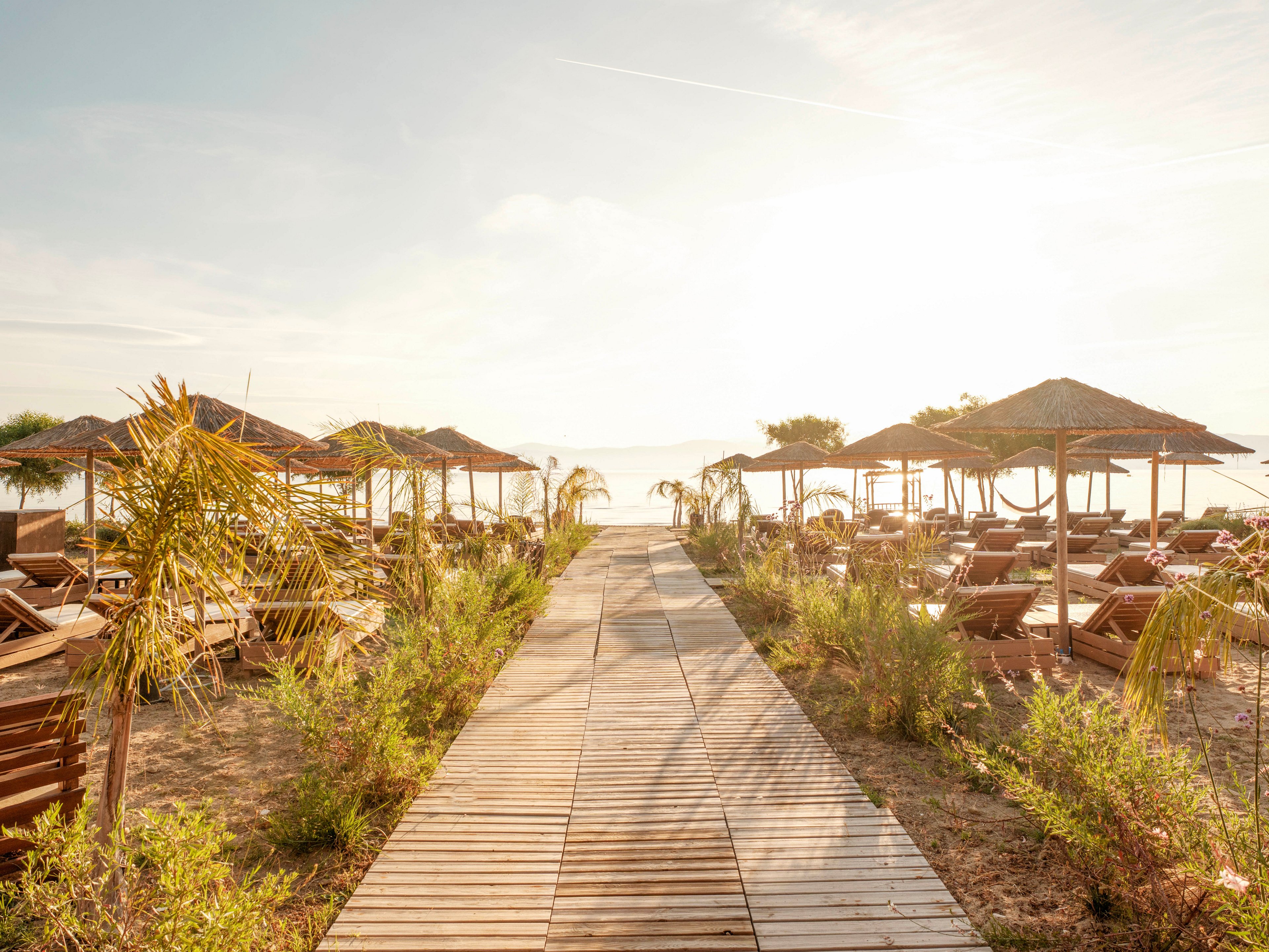 A wooden boardwalk leads through a sandy beach resort lined with lounge chairs and straw parasols. The scene is bathed in warm sunlight, creating a bright and inviting atmosphere. Small palm trees and coastal shrubs frame the pathway on both sides. Calm water and distant hills are visible in the hazy background, suggesting a tranquil seaside destination.