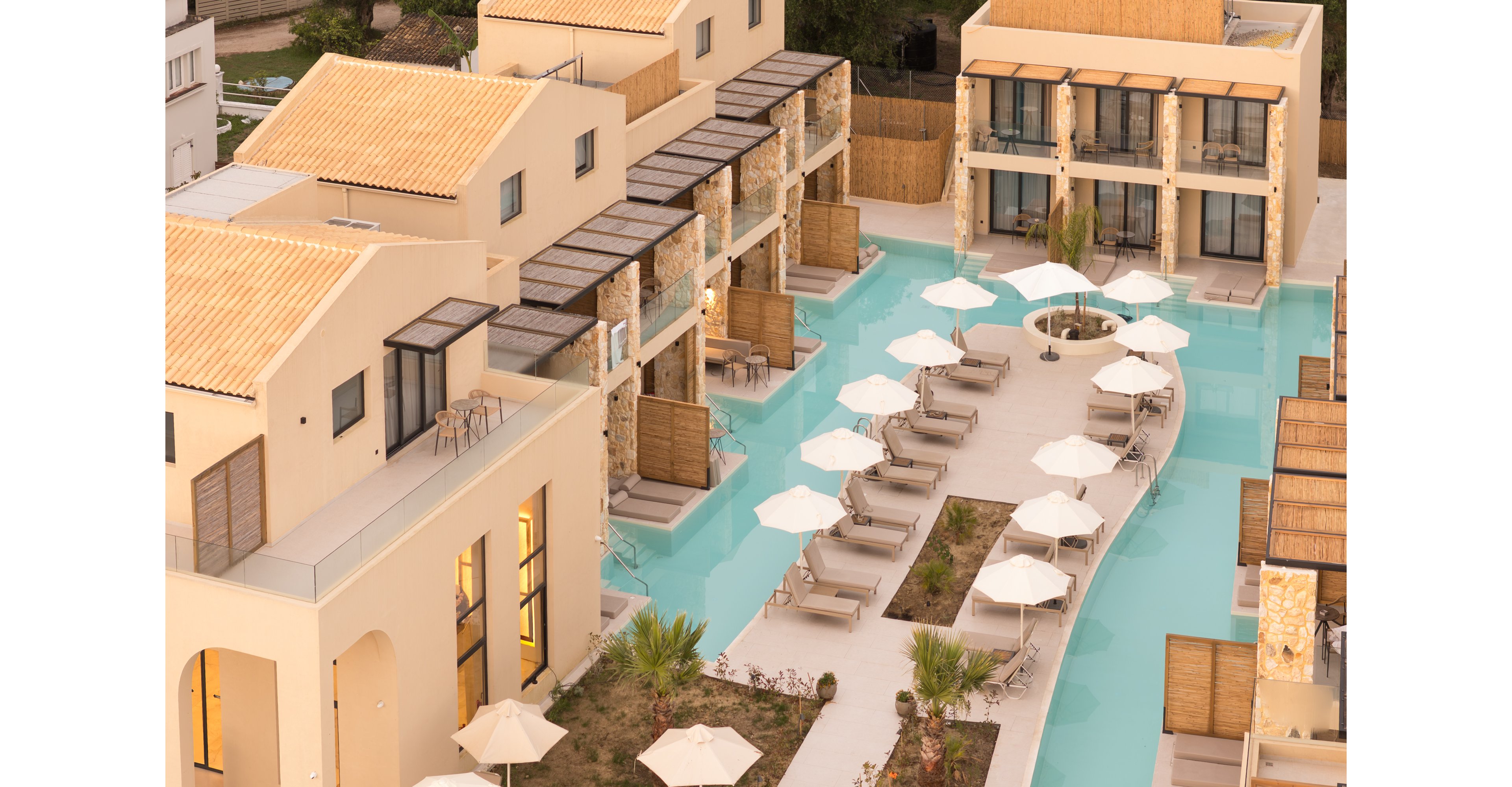 A contemporary resort complex features beige buildings surrounding a narrow turquoise swimming pool. Sun loungers and white umbrellas line the poolside walkway, creating a relaxed vacation atmosphere. The architecture combines clean modern lines with stone accents and wooden privacy screens. Soft evening light enhances the warm color palette and tranquil setting.
