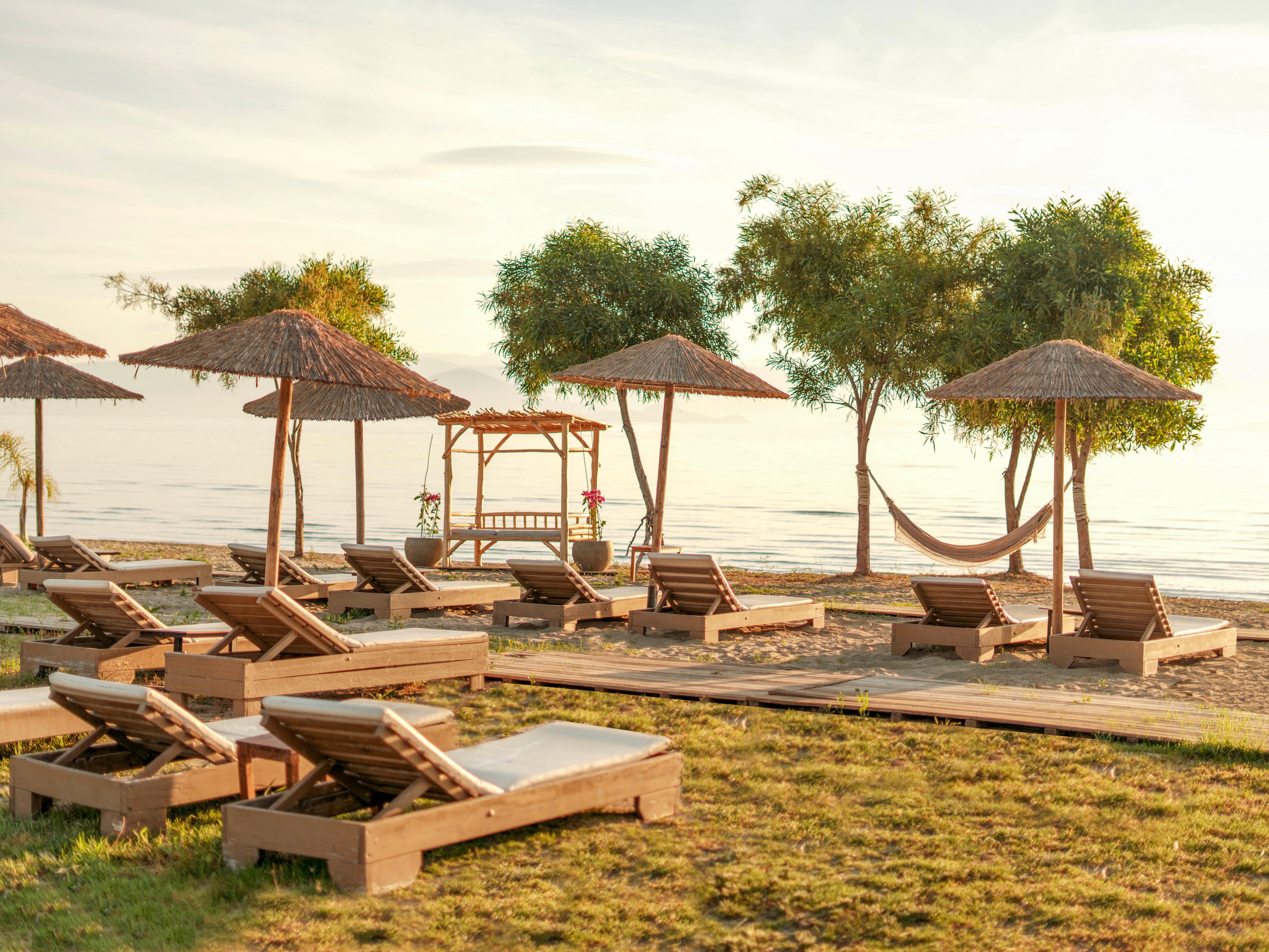 A tranquil beachfront resort features multiple wooden sun loungers arranged on a grassy area facing the calm sea. Straw parasols provide shade, while a wooden gazebo and a hammock hang between trees near the shoreline. The scene is bathed in warm, golden light, suggesting sunrise or sunset. No people are present, emphasizing a peaceful and relaxing atmosphere.
