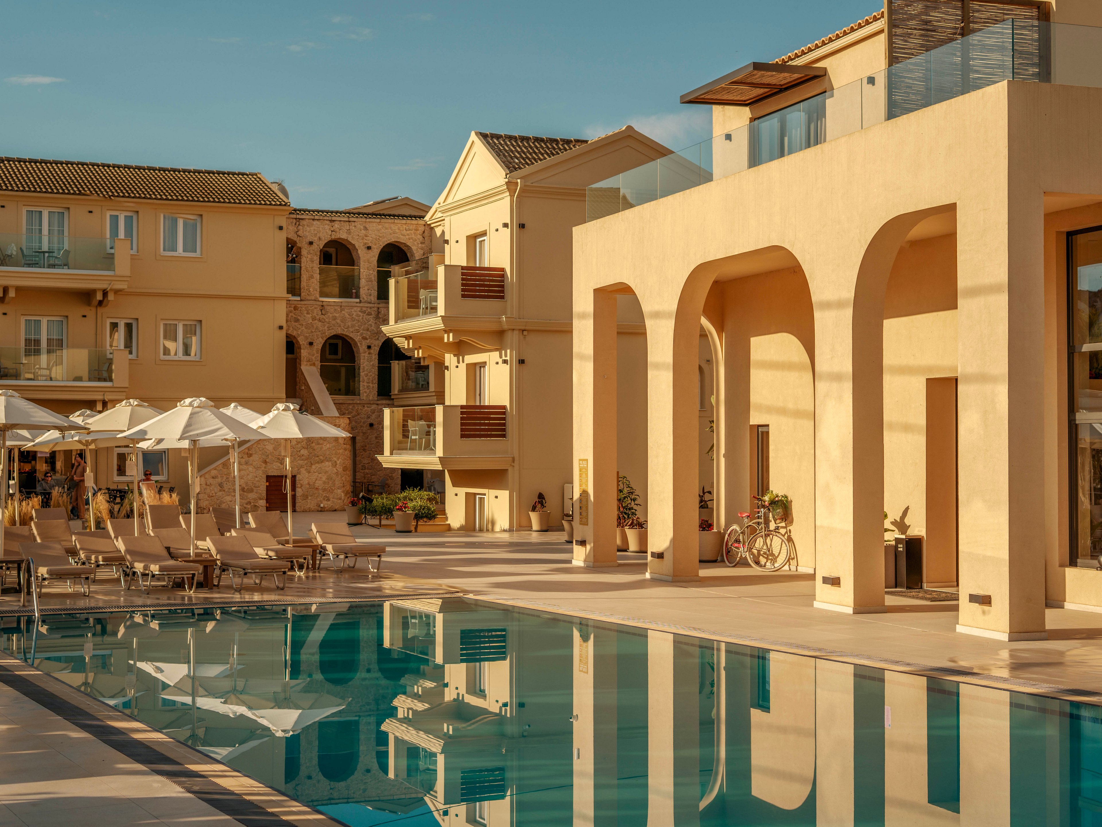 A luxurious resort pool is framed by modern beige buildings and tall arches in warm evening light. Sun loungers and white umbrellas line the poolside, suggesting a relaxing vacation setting. The still water reflects the architecture and sky, enhancing the tranquil atmosphere. A bicycle and potted plants add subtle lifestyle details to the elegant scene.