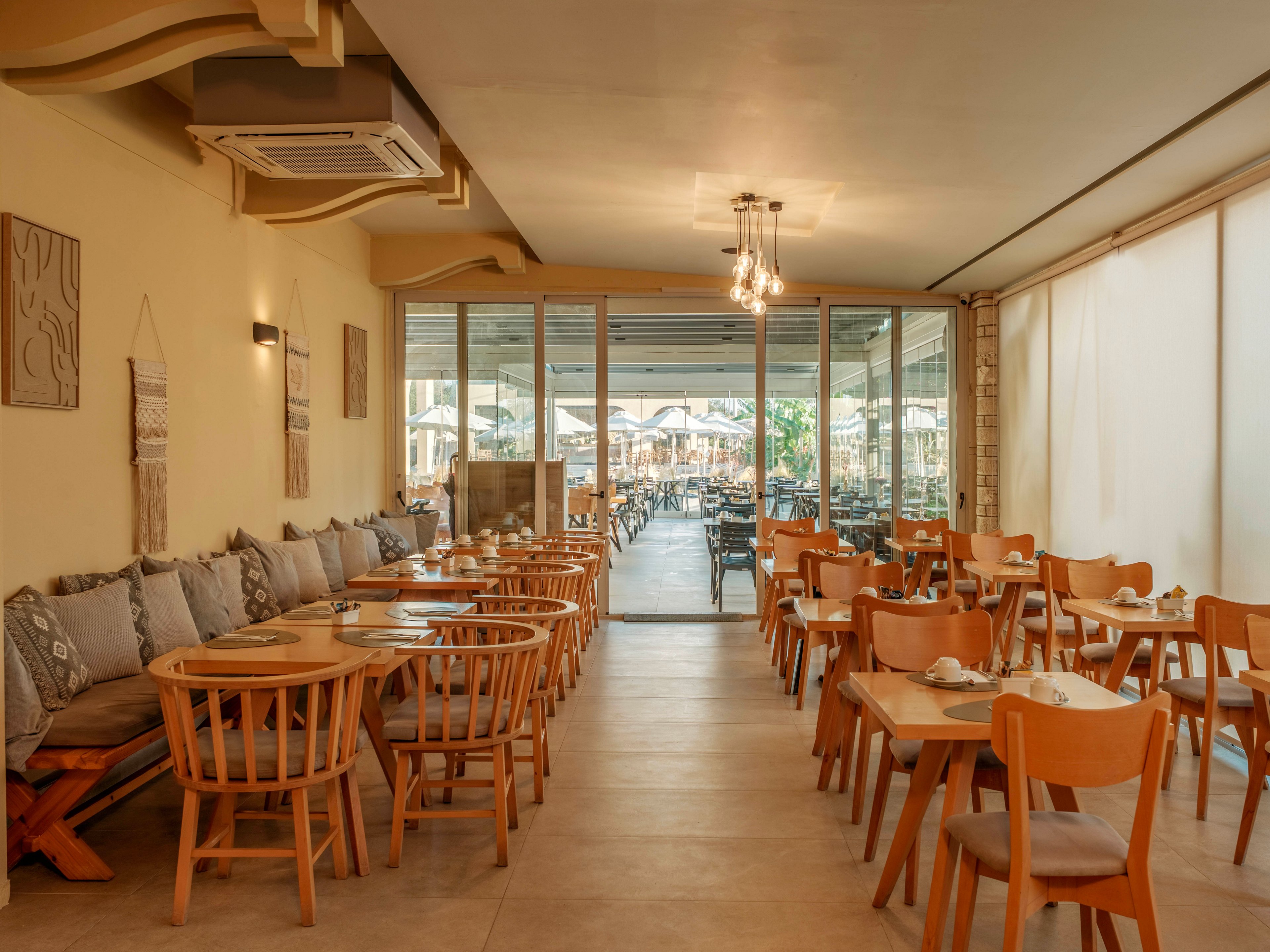 A warm, cozy restaurant interior features wooden tables and chairs neatly arranged in rows. Cushioned bench seating lines one wall, decorated with patterned pillows and simple wall art. Large glass doors open to an outdoor dining area with additional tables and chairs visible in the background. Soft lighting and neutral tones create a relaxed, inviting atmosphere suitable for casual dining.