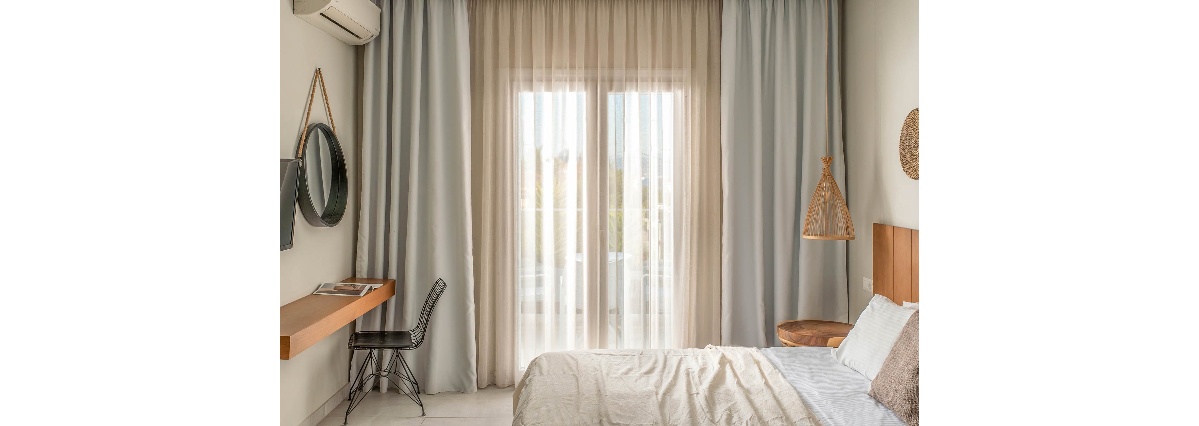 A modern minimalist bedroom features a neatly made bed facing large floor-to-ceiling windows covered with sheer and opaque curtains. Natural light softly filters into the room, illuminating neutral-toned walls and simple furnishings. A floating wooden desk with a chair, wall-mounted TV, and round mirror line one side, while a woven pendant lamp and small side table accent the other. The overall atmosphere is calm, airy, and contemporary.
