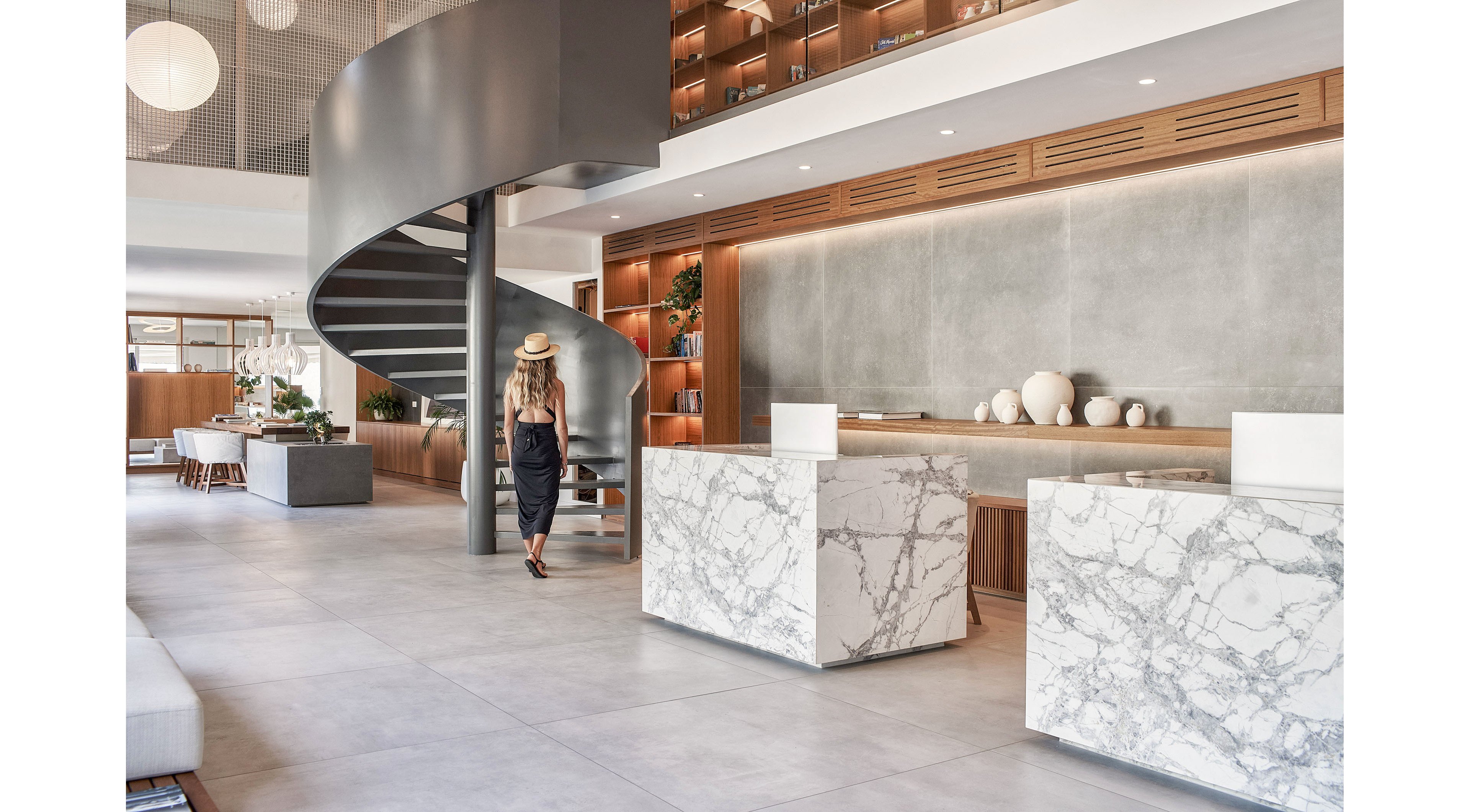 A stylish woman in a black dress and hat walks through a spacious modern lobby. The interior features a dramatic dark spiral staircase, marble reception desks, and large concrete-textured walls. Warm wooden shelving with decorative ceramics and plants adds contrast to the minimalist design. Soft natural light enhances the clean, contemporary atmosphere of the space.